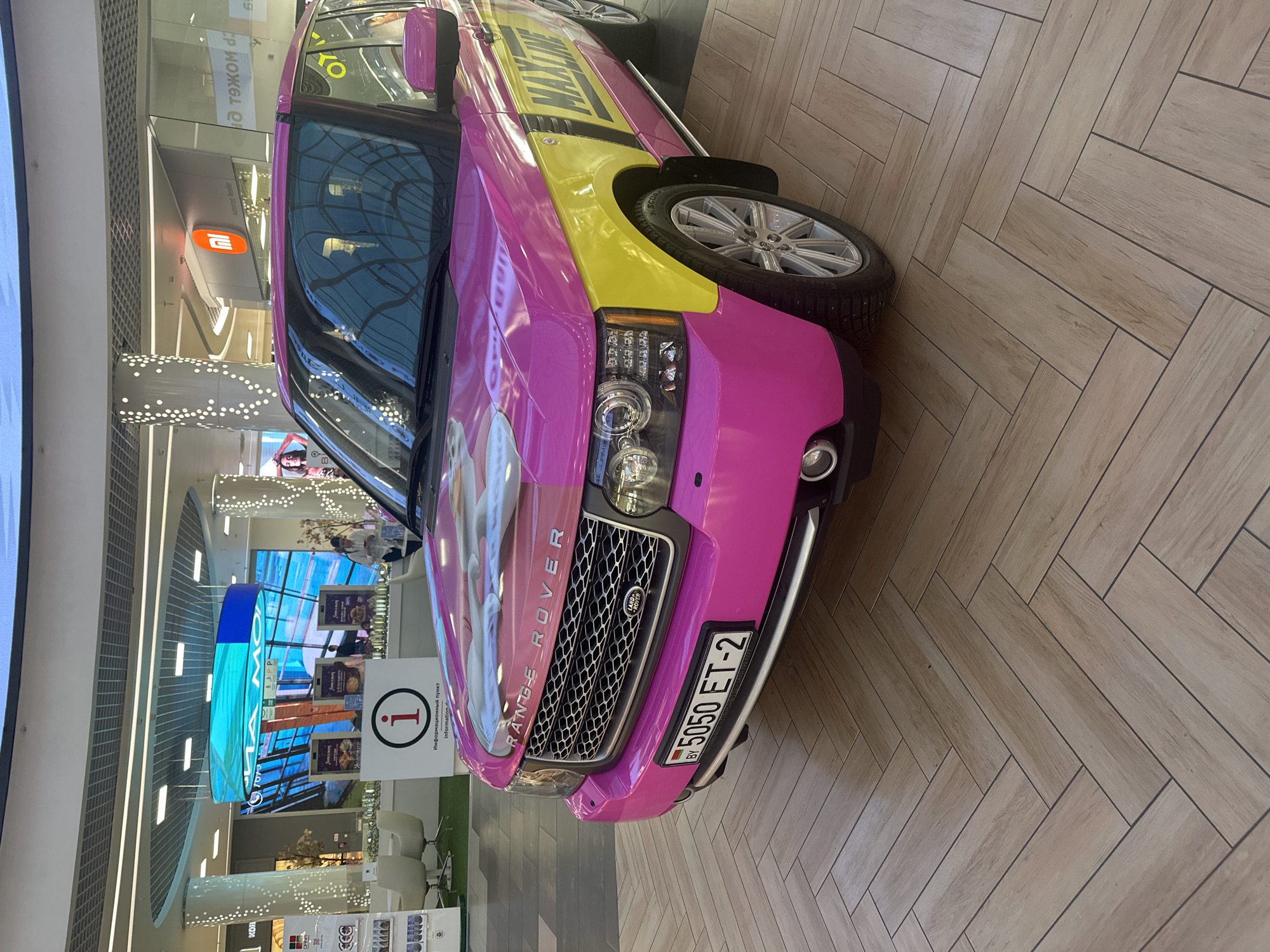 A pink and yellow Range Rover is parked in a shopping mall in Minsk, Belarus. The car has "MAXLINE" written on the side and a license plate that reads "BY 5050 ET-2". In the background, there is a large screen displaying a blue and green image, and an information booth with an "i" logo. There are also some escalators and shops visible. The floor is tiled in a herringbone pattern. There are no people clearly visible interacting with the car.