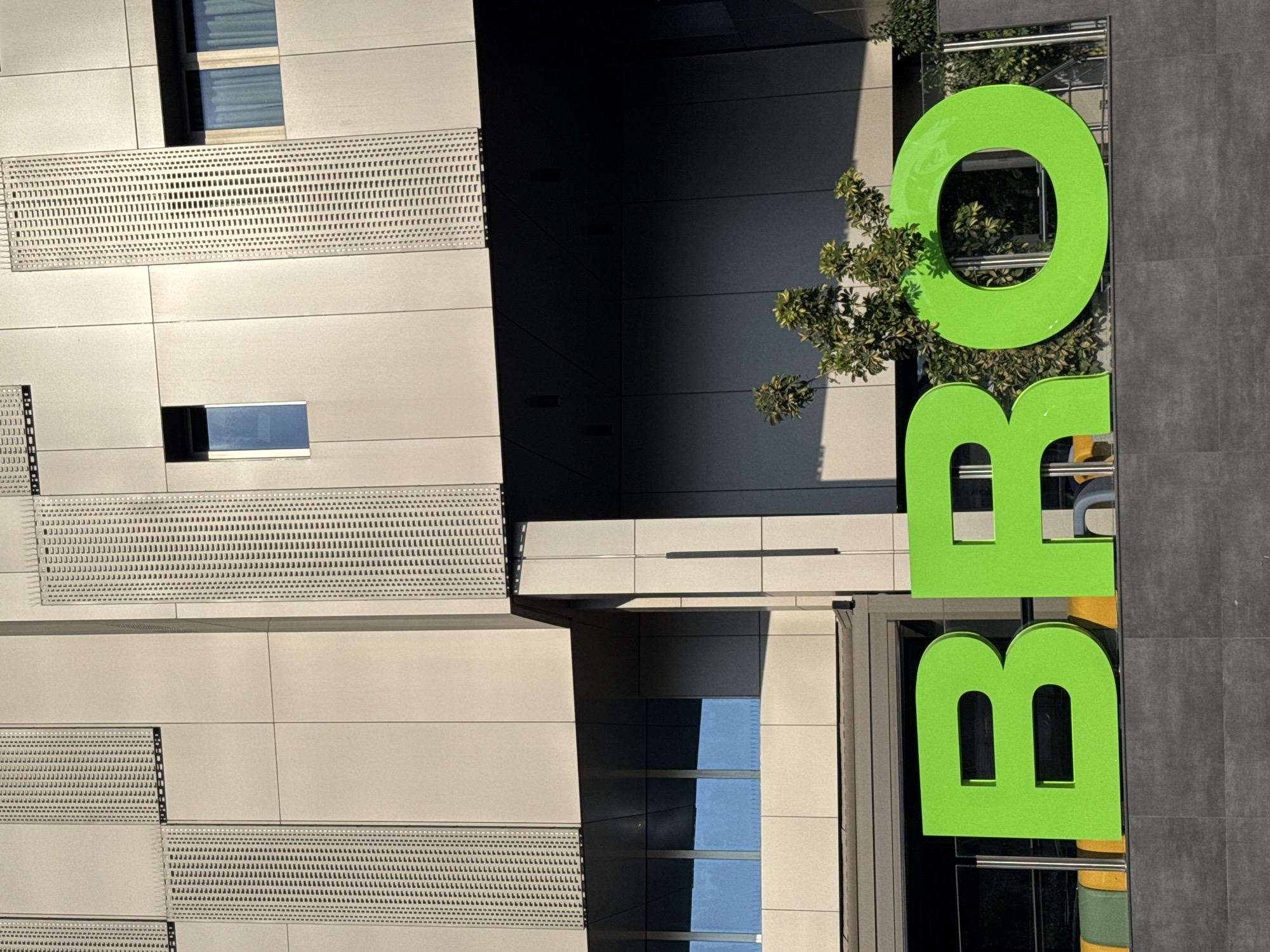 A bright green sign spelling out "BRO" in large, block letters is prominently displayed on a dark gray, textured wall. The sign is located on the facade of a modern building. Above the sign, the building features a light beige exterior with vertical, perforated panels and horizontal window frames. Some windows are visible, showing reflections of the sky and curtains. A small tree with green leaves is growing near the right side of the sign, partially obscuring the "O". The overall scene is a clear, well-lit shot of signage and building architecture. The location is identified as Mesa Geitonia, Cyprus.