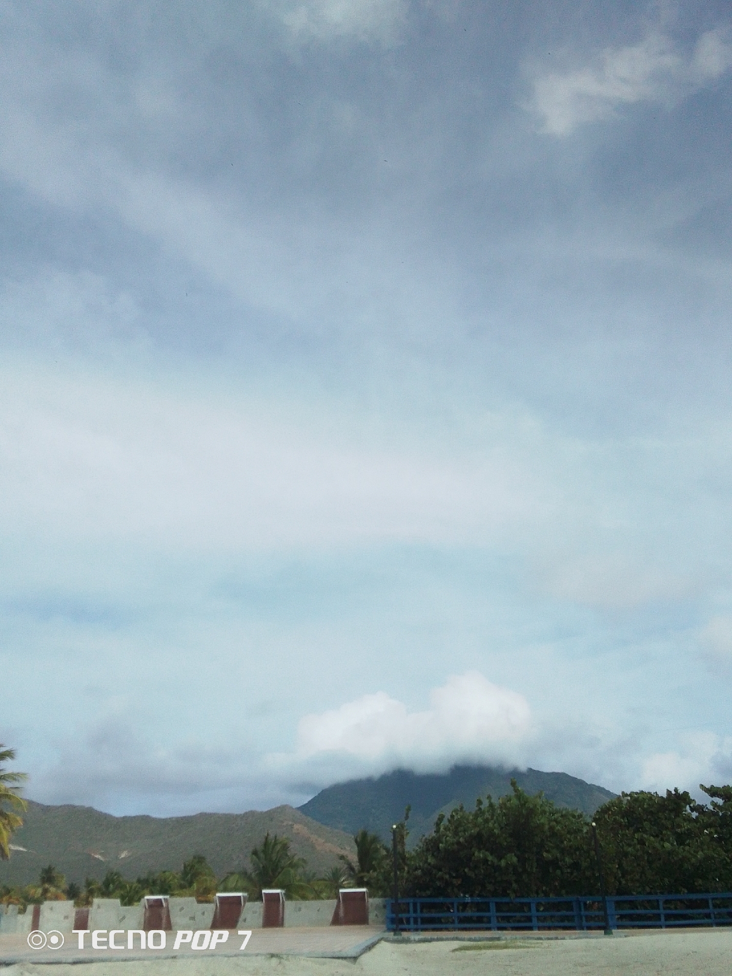 The image shows a landscape view in what appears to be the city of Pedro González, Venezuela. Dominating the scene are mountains covered with vegetation, some obscured by low-hanging clouds. The foreground features a structure, possibly a bridge or building, with a concrete base and a blue railing. Green trees and palms are interspersed in the landscape. The sky is filled with light and dark clouds, suggesting an overcast day. The camera used to take the picture is a Tecno Pop 7, which is indicated by a watermark in the bottom left corner of the image.