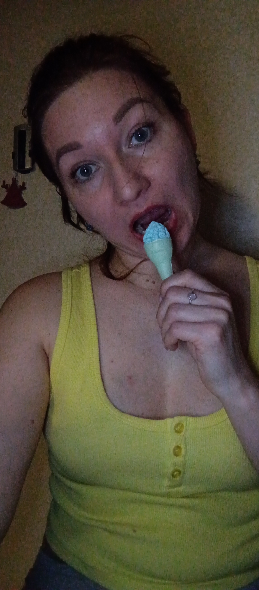 A woman with auburn hair pulled back in a bun is holding a light blue and green ice cream cone to her mouth. She is wearing a yellow tank top with buttons down the front. She has light skin with freckles. She has blue eyes, red lipstick and is wearing a silver ring on her right ring finger. In the background, there is a beige wall with a small silver object and a red decoration hanging on it. The location is unknown.