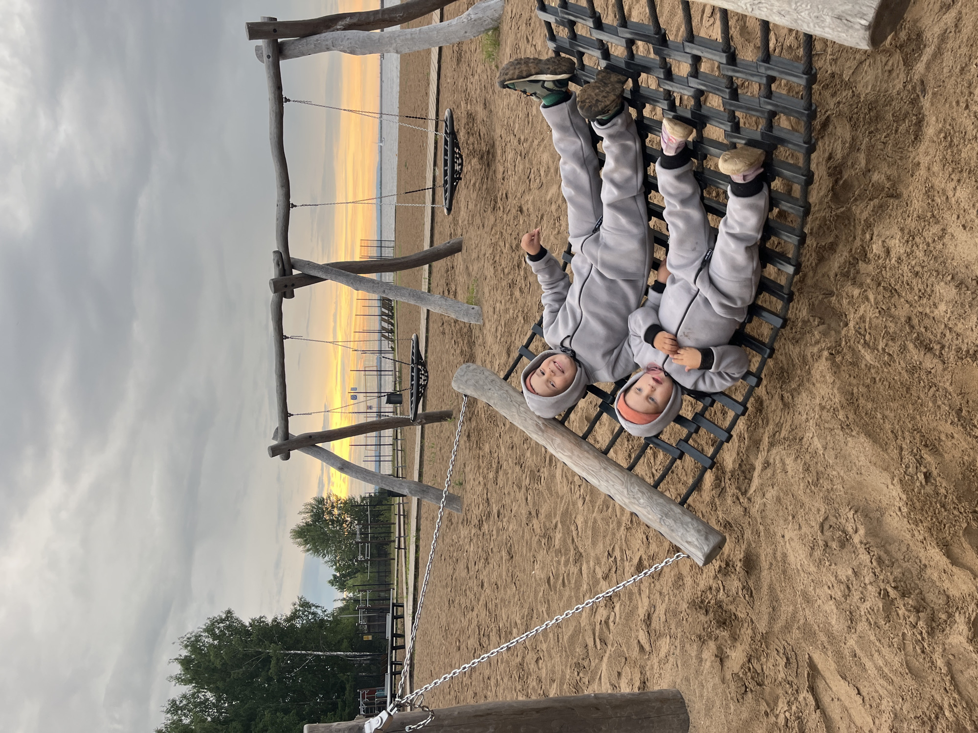 Two young children, a boy and a girl, are lying upside down on a black rope net playground structure. They are both wearing grey hooded jumpsuits. The boy on the left is wearing green and black sneakers, and the girl on the right is wearing pink and beige shoes. The girl has an orange beanie on her head. They appear to be smiling and looking up at the camera.
The playground is situated on a sandy surface, with a wooden swing set visible in the background. Beyond the swings, there's a body of water that appears to be a river or lake. The sky is overcast with the soft glow of sunset, casting a warm light on the horizon. Trees and other playground equipment are visible in the middle ground. The overall scene suggests a relaxed and playful atmosphere at a park or beach area in Chaikovsky, Russia, likely during the late afternoon or early evening. There is no visible text in the image.