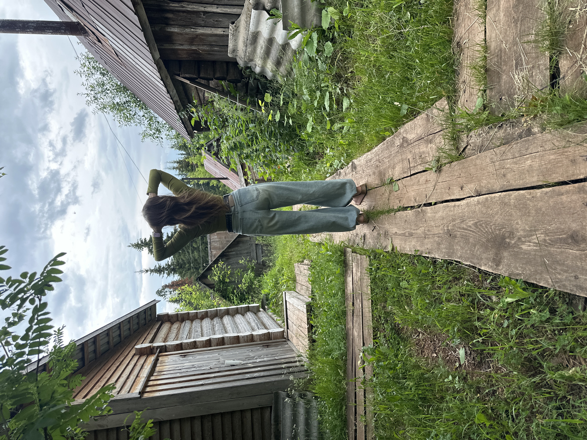 In the city of Ilyinsky, Russia, a woman stands on a wooden walkway, her back to the camera. She is wearing a long-sleeved green shirt and loose-fitting light blue jeans. Her hands are clasped behind her head, and her hair is long and brown. The walkway is made of weathered wooden planks and is surrounded by lush green grass and foliage. To the left and right of the walkway are rustic wooden structures, appearing to be small cabins or sheds. The sky above is overcast with dramatic clouds. In the background, more trees and structures are visible, suggesting a rural or semi-rural setting.