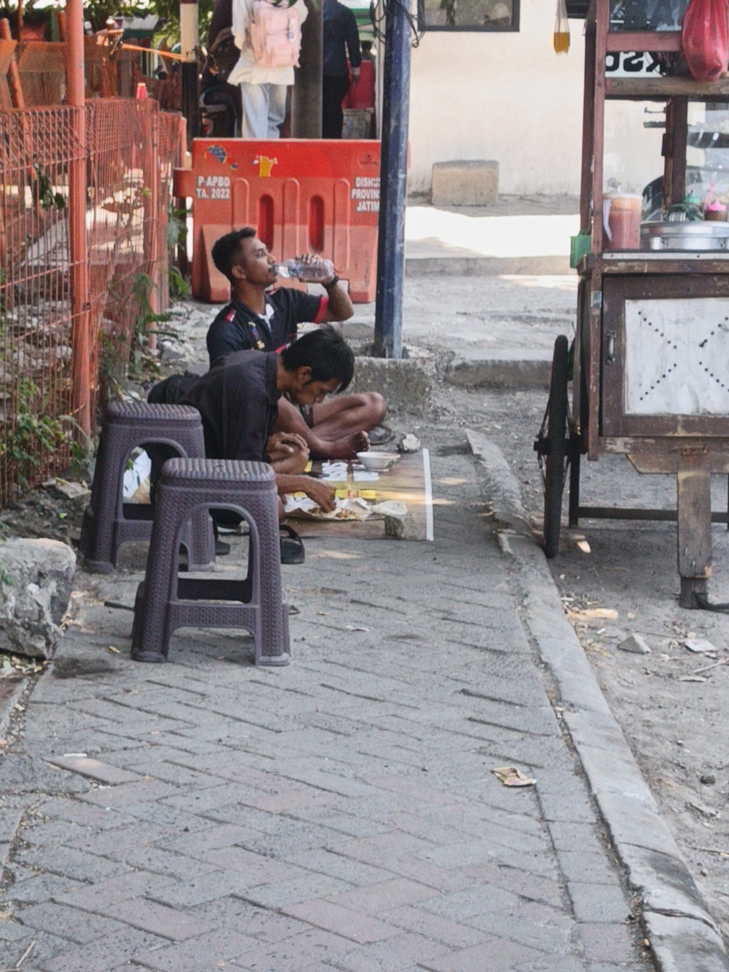In Kedungrejo, Indonesia, two men are seen seated on the ground next to a street. The man on the left is wearing a dark shirt with red trim and is drinking from a plastic bottle. The man on the right, wearing a dark shirt, is leaning forward and appears to be eating from a plate on the ground. They are positioned near two brown plastic stools. In the background, a food cart with a wooden frame and metal shelves is visible, with a red bag hanging from it. The street is paved with brick. A red metal fence runs along the left side of the scene, with a construction barrier featuring text and a globe graphic behind it. A blue pole stands near the barrier. The environment is outdoors, under daylight.
