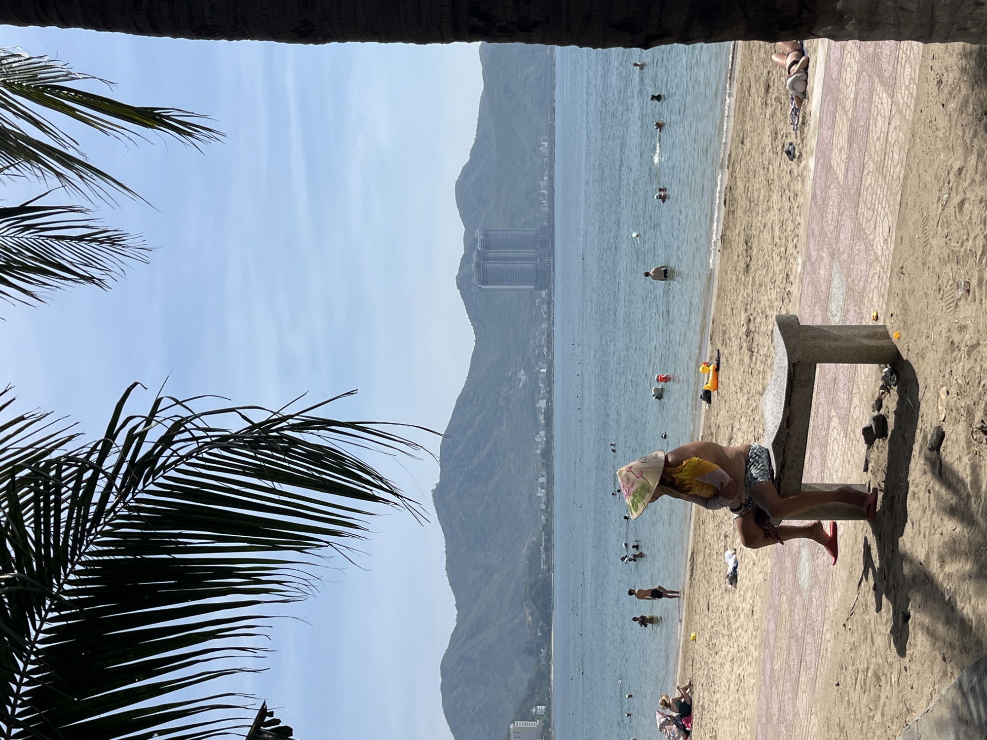 A wide shot captures a serene beach scene in Bac Nha Trang Ward, Vietnam, under a bright, clear sky. In the foreground, a palm frond partially obscures the view, casting dappled shadows on the sandy ground and a concrete bench. A person sits on the bench, facing the ocean, wearing a conical hat and patterned shorts, with red sandals on their feet. They hold a yellow bag in their lap.
The beach extends towards the water, where several people are enjoying the ocean. Some are swimming, while others stand in the shallows. A yellow inflatable toy is visible near the shore.
In the mid-ground, the calm sea stretches out, reflecting the sky. The background is dominated by a range of lush, green mountains that line the coast, with a city visible nestled at their base. Two tall, modern buildings stand out prominently against the mountainside, hinting at the urban development in the area. The overall atmosphere is peaceful and inviting, suggesting a pleasant day at the beach.
