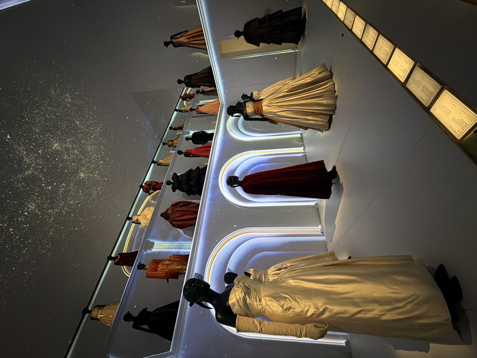 The image displays an interior view of a fashion exhibition, identified as the Galerie Dior at 30 Avenue Montaigne in Paris, France. The scene features a dramatically lit room with a dark ceiling adorned with numerous small, bright points of light, creating the impression of a starry night sky.
Multiple black, featureless mannequins are positioned on various display levels and on the floor, showcasing an extensive collection of haute couture gowns and elaborate garments. In the foreground, to the left, a mannequin wears a floor-length, strapless champagne satin ball gown with a draped bodice, long matching gloves, and a black veiled headpiece. To its right, another mannequin is draped in a floor-length, deep red cape. Further right, a mannequin displays a strapless, champagne-colored ball gown with an orange sash, adjacent to another mannequin in a dark, multi-tiered full-skirted gown.
The midground and background feature a wall characterized by a series of tall, illuminated arched niches, outlined by glowing white light. Within these arches and on shelves above them, approximately 15-20 additional mannequins are visible, exhibiting formal gowns and elaborate ensembles in colors such as black, gold, orange, various reds, and beige.
The floor is a uniform, light color. Along the floor in the lower right corner, a linear arrangement of small, illuminated square plaques with text provides informational captions for the displayed items. No human figures are present; the scene is static, focused on the fashion display.