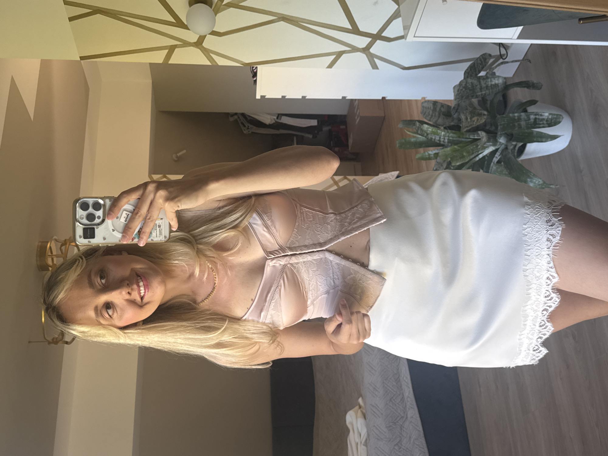This is a vertically oriented, eye-level, full-body mirror selfie of a young woman with long blonde hair. She is smiling and looking at her reflection, holding a smartphone in her left hand. She is wearing a light pink, lace-trimmed bra and a white, lace-trimmed slip. The background appears to be a bedroom or dressing area, with a dresser and a potted plant visible to the right. A geometric-patterned ceiling design is also visible in the reflection. The lighting suggests it is daytime.