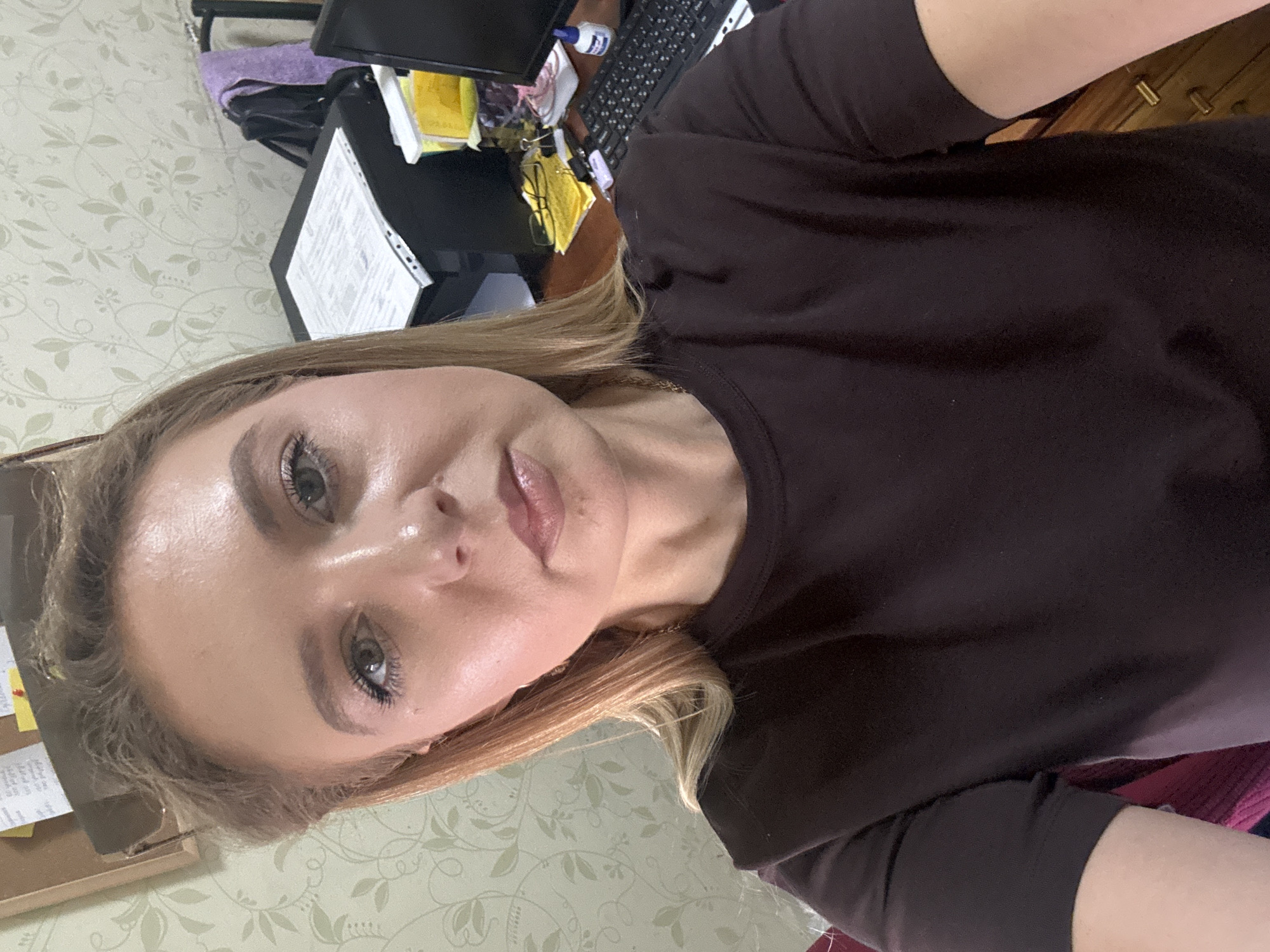 A young woman with blonde hair pulled back, wearing a dark brown t-shirt, is taking a selfie in a room. She has on light makeup and is looking directly at the camera. Behind her, a desk is cluttered with various items including papers, a computer monitor, a keyboard, a small bottle, and a purple cloth draped over a chair. The wall behind the desk has a light green floral wallpaper. The scene appears to be an indoor office or personal workspace.