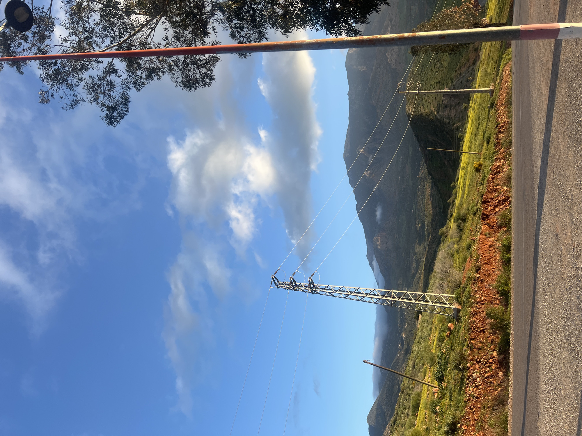 The image depicts a landscape in Ouaouizert, Morocco, featuring a partly cloudy sky, mountains, and power lines. A metal utility pole stands prominently in the scene. A paved road is visible on the right side of the image, with a reddish-brown dirt area adjacent to it. Vegetation covers the mountain slopes. A red and white painted barrier is partially visible at the top of the image. The mountain in the background is partially obscured by clouds. The image appears to be taken at an angle, with the horizon tilting towards the right.