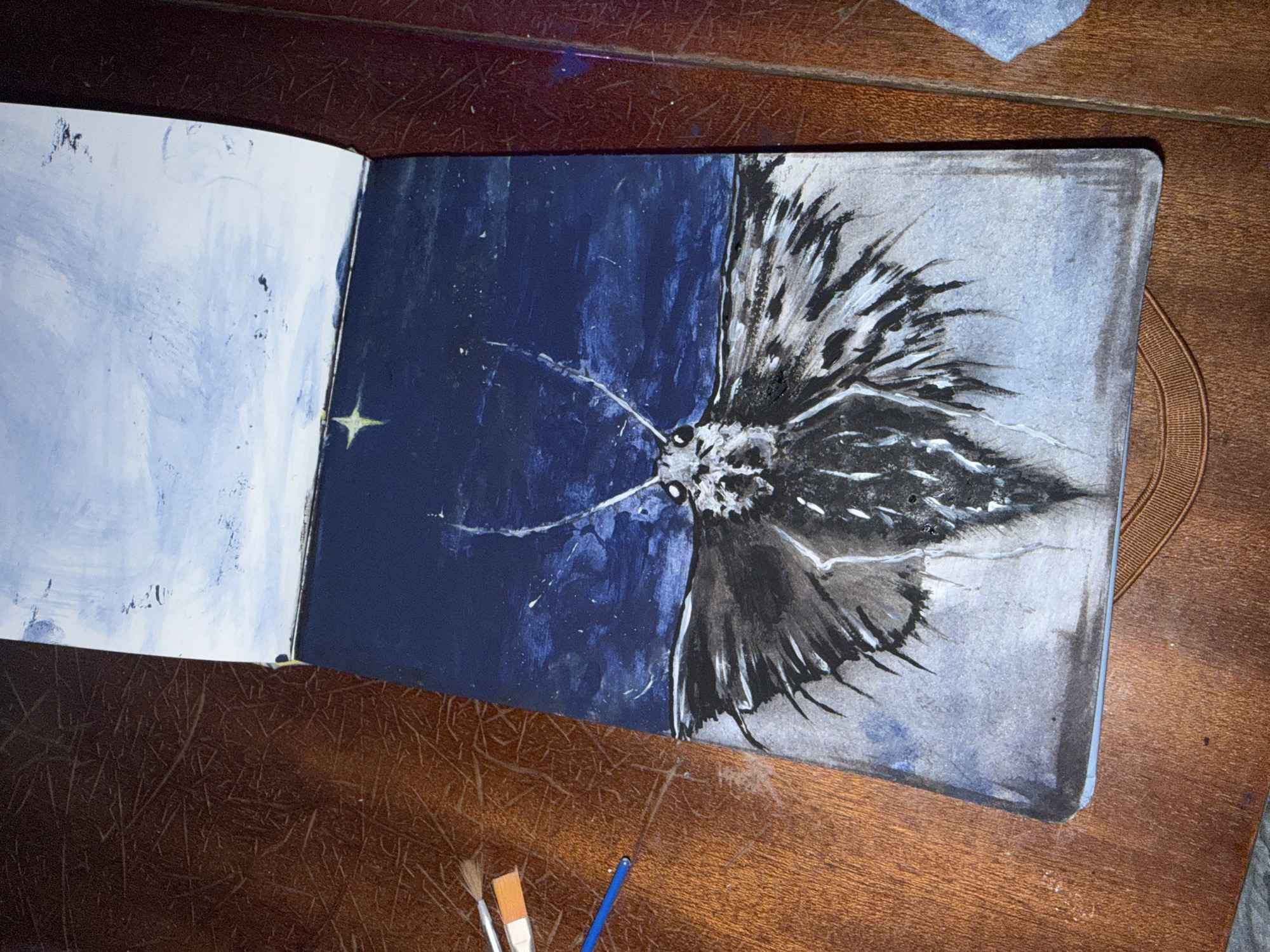 An open sketchbook lies on a wooden surface, revealing a two-page painting. The right-hand page displays a grayscale image of a moth, wings spread, with a detailed head and body. The wing tips fade into a lighter gray background. The left-hand page features a deep blue background with a single, small yellow star. The remaining area is a lighter blue with scattered paint splatters. Three paintbrushes are visible near the sketchbook. The wooden surface beneath the sketchbook appears to be a table. Location information is not available in the provided image.