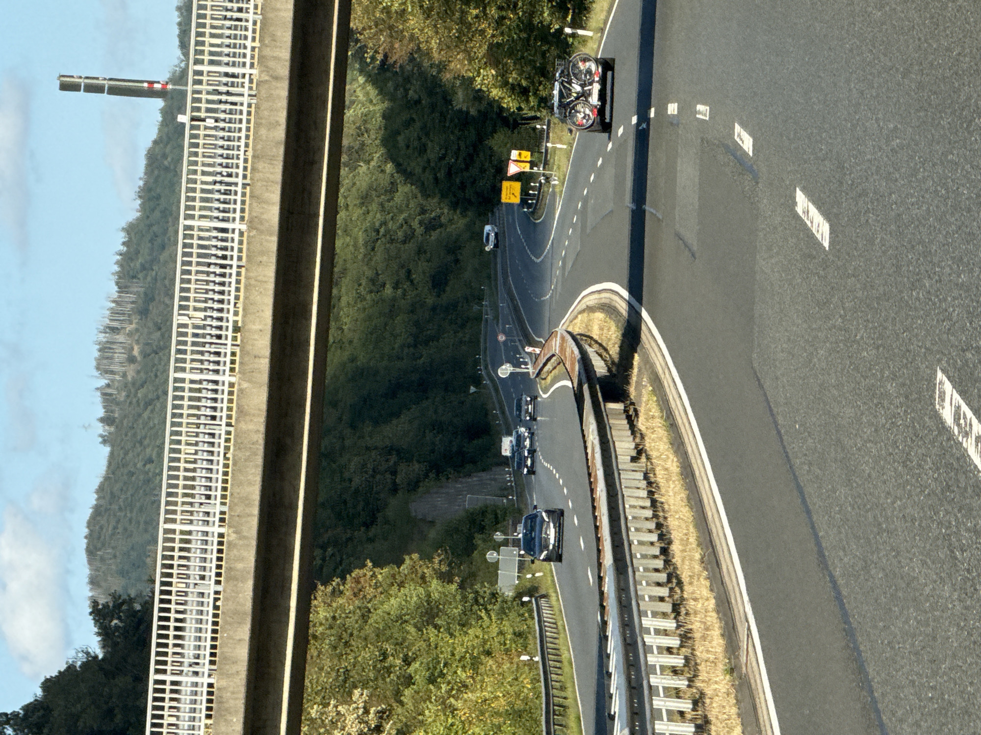 The image depicts a view from a vehicle traveling on a highway under a bridge, with the city of Bad Lauterberg im Harz, Germany, visible in the background. The highway has multiple lanes and is surrounded by lush green trees and hills. Several cars are driving on the highway, some of which are in the foreground. One car in the right lane has two bicycles mounted on a rack at the back. The bridge overhead is a concrete structure with a white metal railing. The sky is blue with some scattered clouds. The overall scene suggests a sunny day on a scenic drive through the German countryside.