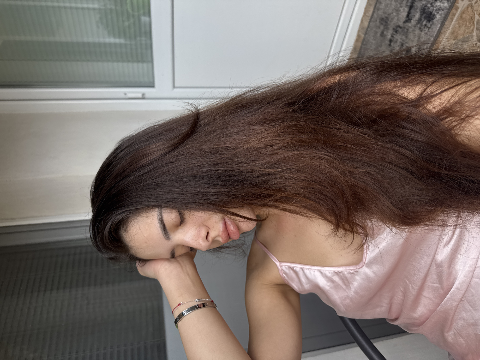 The image displays a female individual in a medium-close shot, seated on an outdoor balcony in Plovdiv, Bulgaria. She is positioned with her head resting on her right hand, facing towards the right side of the frame, and her eyes are closed, indicating a moment of repose. Her long, dark brown hair, exhibiting layers and subtle waves, cascades over her left shoulder and down her back. Her skin is fair, with visible freckles across her nose and upper cheeks. She has full, pink-toned lips. She is wearing a light pink, satin-like camisole with thin shoulder straps. On her right wrist, she wears three bracelets: a wide silver-colored band bracelet with screw motifs, a delicate silver-colored chain bracelet, and a thin red string bracelet. The background features a dark, curved element on the left, likely part of a chair or railing. Behind her, a white-framed glass door or large window with interior horizontal blinds is visible, suggesting an adjacent indoor space. The balcony floor is tiled, and a dark, patterned mat is visible in the lower right section of the frame. The scene is illuminated by soft, ambient daylight.