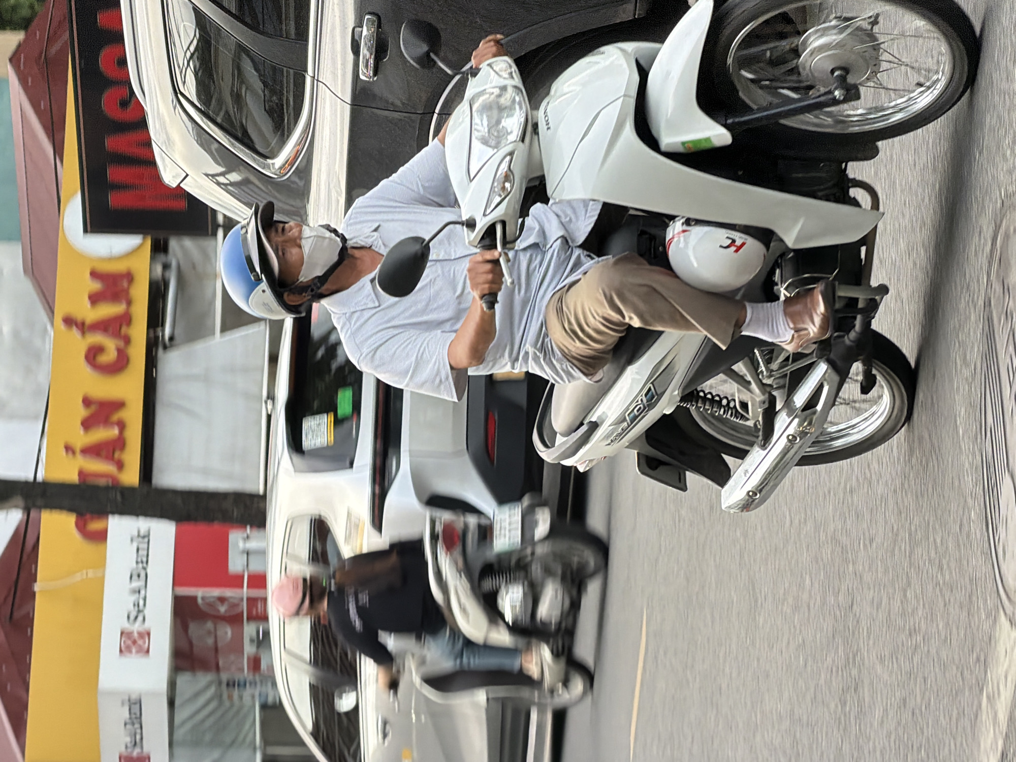 In Thuận Hóa Ward, Vietnam, a middle-aged man wearing a blue and white helmet and a white face mask rides a white Honda scooter down a street. He is wearing a light blue collared shirt, khaki pants, white socks, and brown loafers. The scooter has a white body with red and black accents. In the background, a dark SUV is parked or moving slowly, and other vehicles and buildings are visible. The scooter is in motion, indicated by the blurred background and the dynamic pose of the rider.