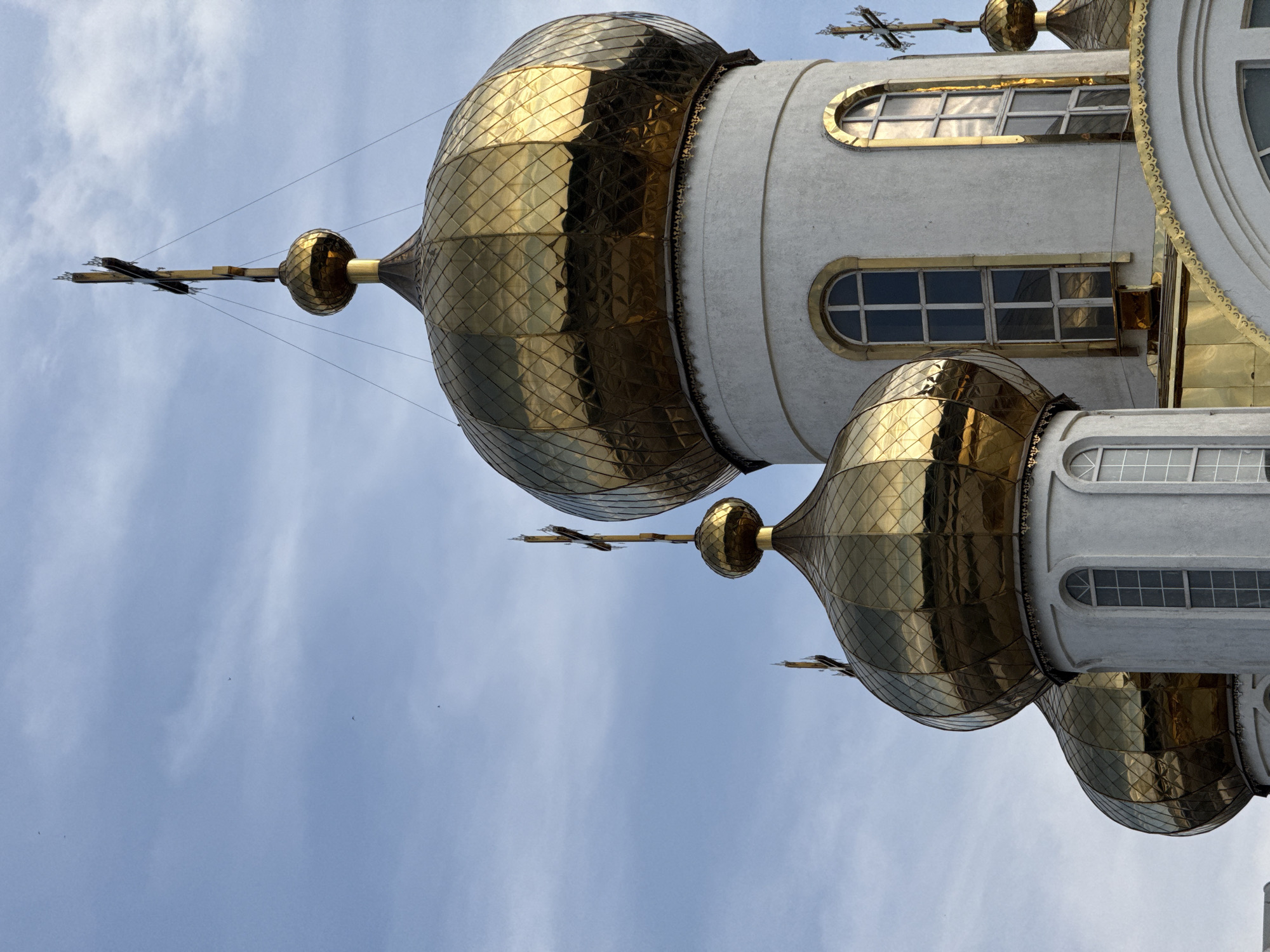 A close-up, eye-level, medium shot captures the golden domes and white walls of an Orthodox church against a light blue sky with wispy clouds. Several golden domes, each topped with a cross, are visible. The largest dome in the foreground reflects the sky and surrounding architecture. White cylindrical towers support the domes, featuring arched windows with multiple panes and golden trim. The image is sharply focused, emphasizing the textures and details of the building. Several tiny dark specks, likely birds, are scattered across the sky. The scene is devoid of any people or immediate human activity.
