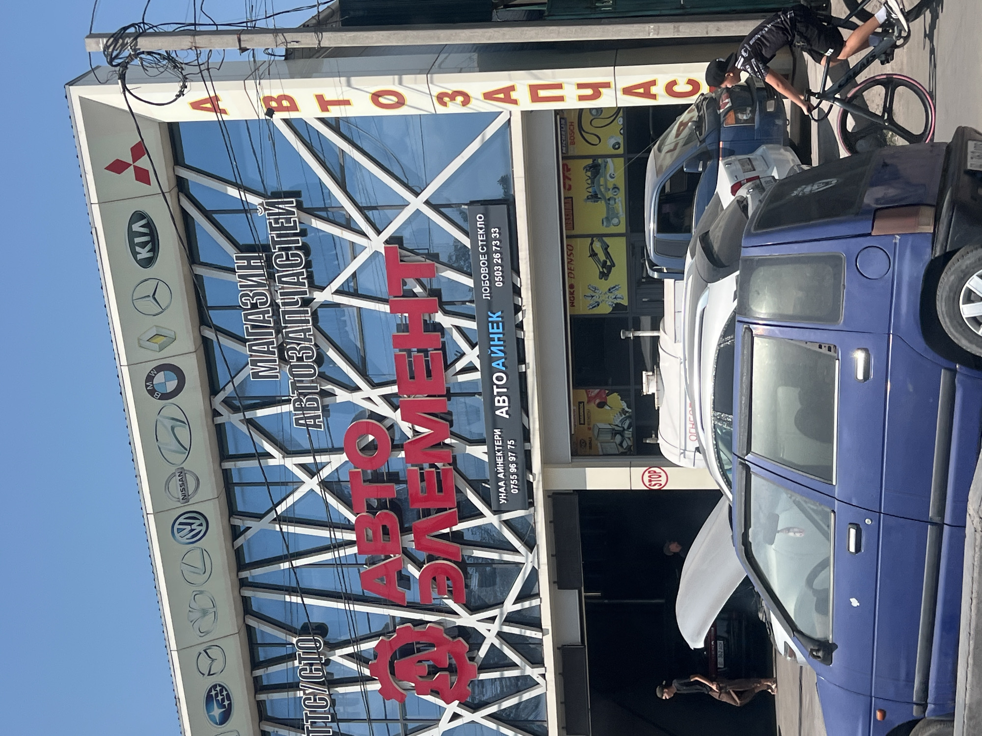 In Bishkek, Kyrgyzstan, a storefront for an auto parts store is prominently displayed, featuring large red lettering that reads "АВТО ЭЛЕМЕНТ". Above this, the store is identified as "МАГАЗИН АВТОЗАПЧАСТЕЙ". The building's facade is characterized by a modern glass design with white diagonal accents. Along the top edge of the building, various car manufacturer logos are visible, including Subaru, Mazda, Honda, Volkswagen, Nissan, Hyundai, BMW, Renault, Mercedes-Benz, Kia, and Mitsubishi.
To the right of the store's name, a vertical sign in yellow with red lettering spells out "АВТОЗАПЧАСТИ". Below the main signage, a smaller black sign advertises "ЛОБОВОЕ СТЕКЛО" (windshield) with a phone number. In front of the store, several vehicles are present, including a blue sedan in the foreground, a white hatchback, and a blue pickup truck. Two men are visible near the entrance of the store, one standing by a car with its hood open, and another further inside the shaded area. A young person is riding a bicycle on the sidewalk in front of the store, moving from right to left. A red "STOP" sign is mounted on a pole near the entrance. The environment appears to be a commercial area with overhead power lines.