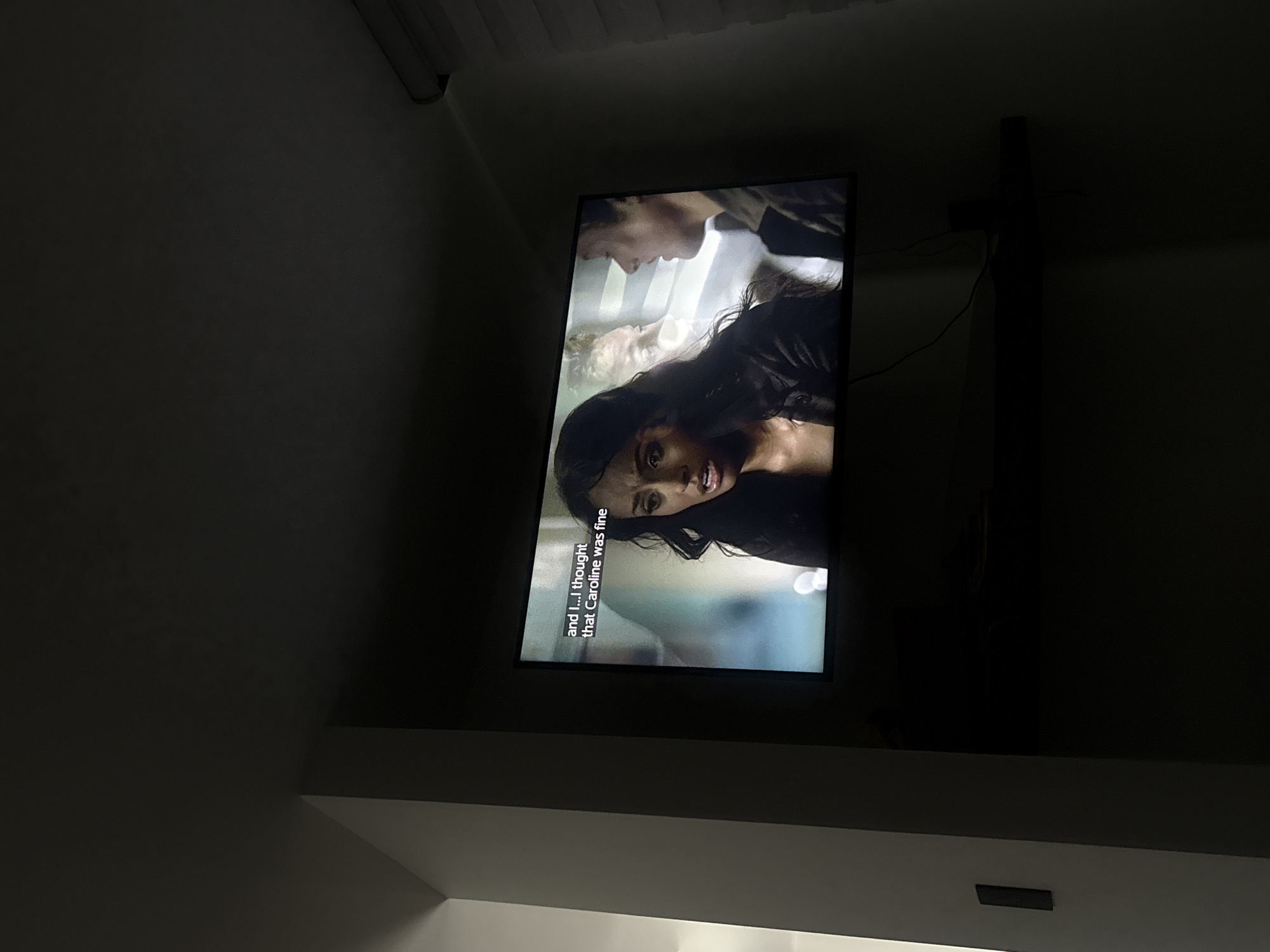 A television mounted on a wall displays a scene from a show with subtitles. The scene includes a dark-haired woman in the foreground and a man looking down in the background. There is also a fair-haired woman who appears to be deceased. The setting is dark, and the image is taken at an angle. The location is not evident in the image.
