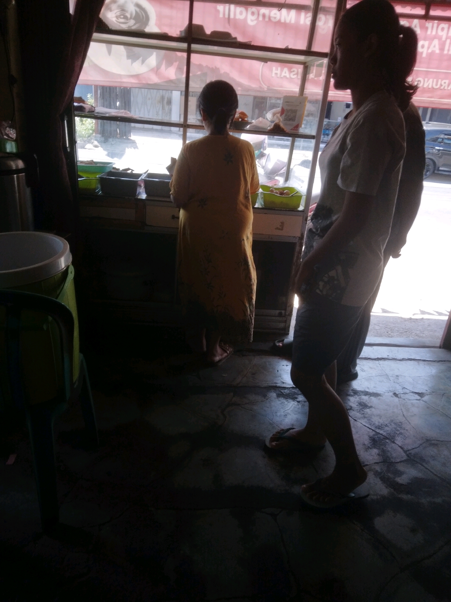 The image depicts an interior scene, likely a small shop or food stall, located in Purwokerto, Indonesia. The setting is dimly lit, with strong natural light entering from a large window or opening at the rear. In the foreground, a person's legs and feet are visible, wearing flip-flops and standing on a wet, cracked concrete floor.
Behind this individual, a woman is visible from the back, wearing a yellow patterned dress. She is standing near a counter or display area, which contains several containers, one appearing to be a green plastic tub filled with items, possibly food. To the left, a large cylindrical container, possibly a water dispenser or trash bin, is partially visible, along with a dark green chair.
The background reveals a shop interior with shelves and a counter. Through the large opening, a street scene is faintly discernible, with a parked car and buildings. A banner with red and white text is mounted above the opening. The overall impression is one of a simple, working establishment.