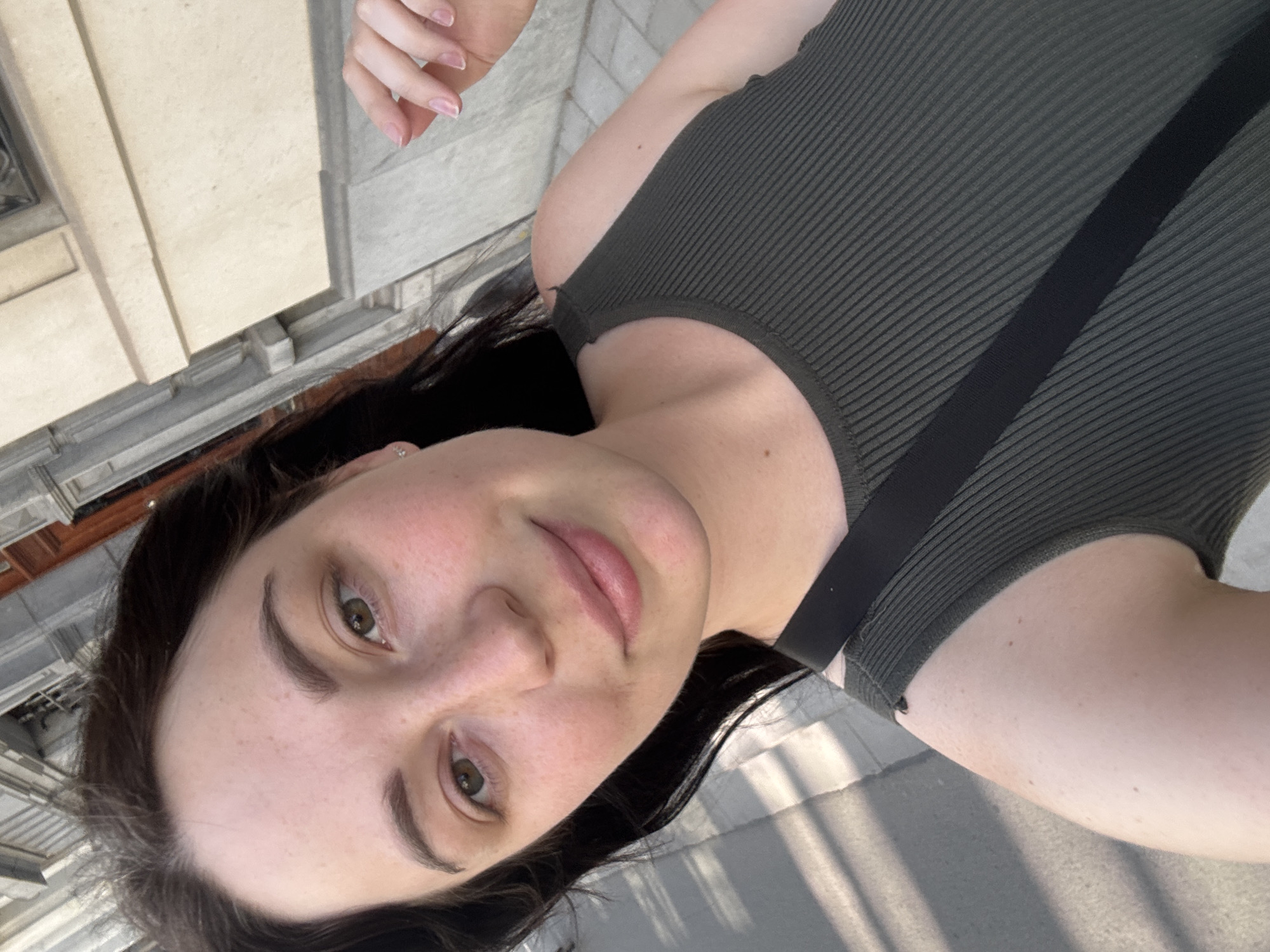 A close-up, selfie-style photograph shows a woman with dark hair, fair skin, and freckles. She is wearing a dark olive-green ribbed tank top. Her right hand is visible in the upper right corner of the image, with manicured nails. The background suggests an outdoor urban setting with a building facade and a sidewalk. The lighting indicates it is daytime, possibly late afternoon due to the shadows. The specific location in Antwerp, Belgium is not discernible from the visual information provided.