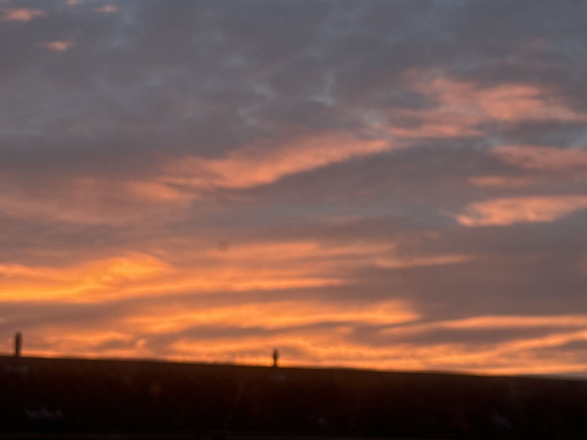 The image captures a colorful sunset in Great Yarmouth, United Kingdom. The sky is filled with streaks of orange and grey clouds. Silhouetted against the bright sky is what appears to be a rooftop, with two small, indistinct figures standing on it. The lower portion of the image is dominated by the dark shape of the roofline.