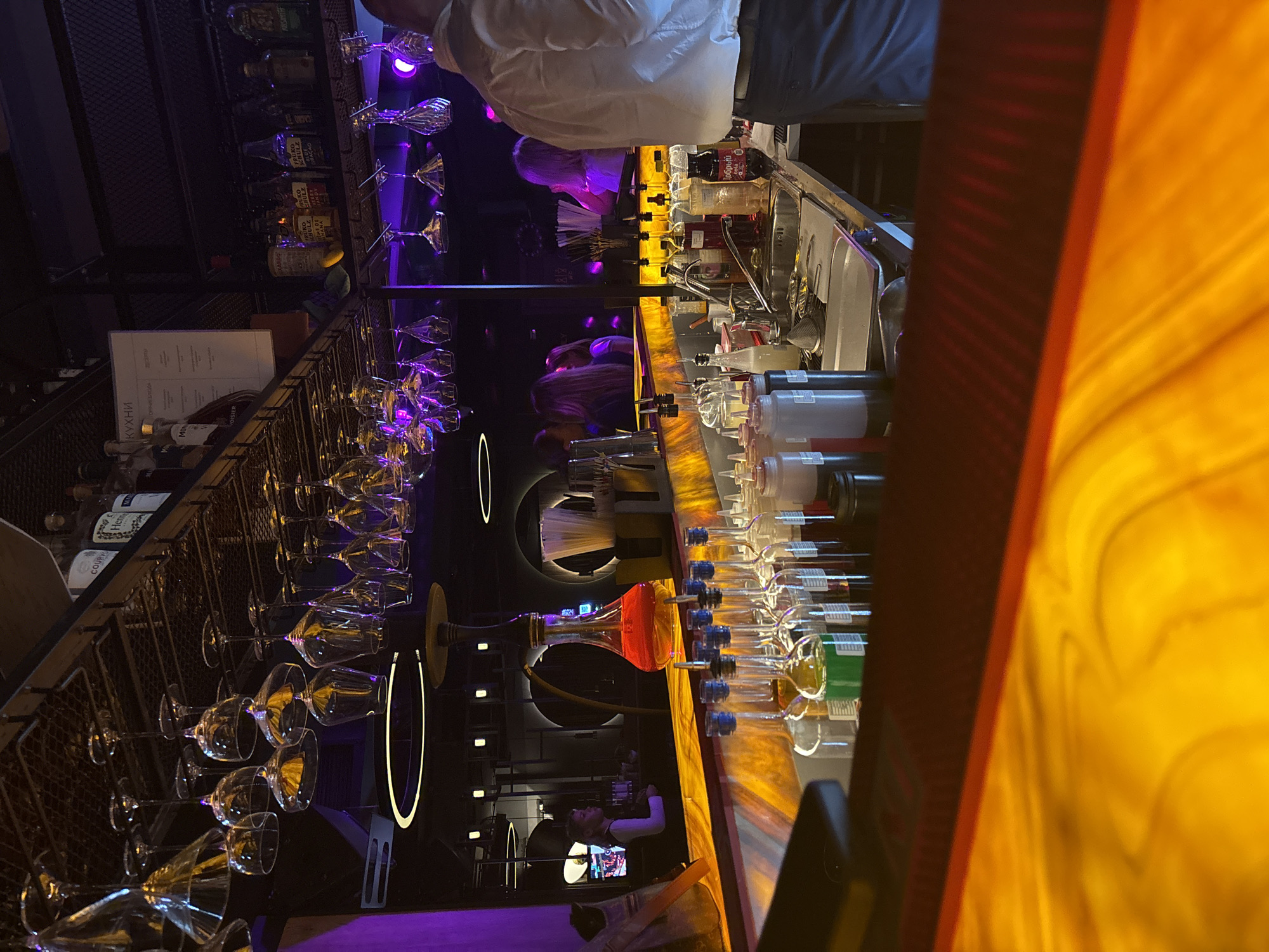 The image captures the interior of a bar, likely in Perm, Russia, based on text on the menu. The bar is well-lit and features a long countertop, lined with numerous liquor bottles and dispensing containers. Several empty wine glasses hang upside down in metal racks above the counter. A bartender is present, visible in the background, near a monitor displaying content. Decorative lighting fixtures add ambiance to the space. A person in a white shirt stands behind the counter.