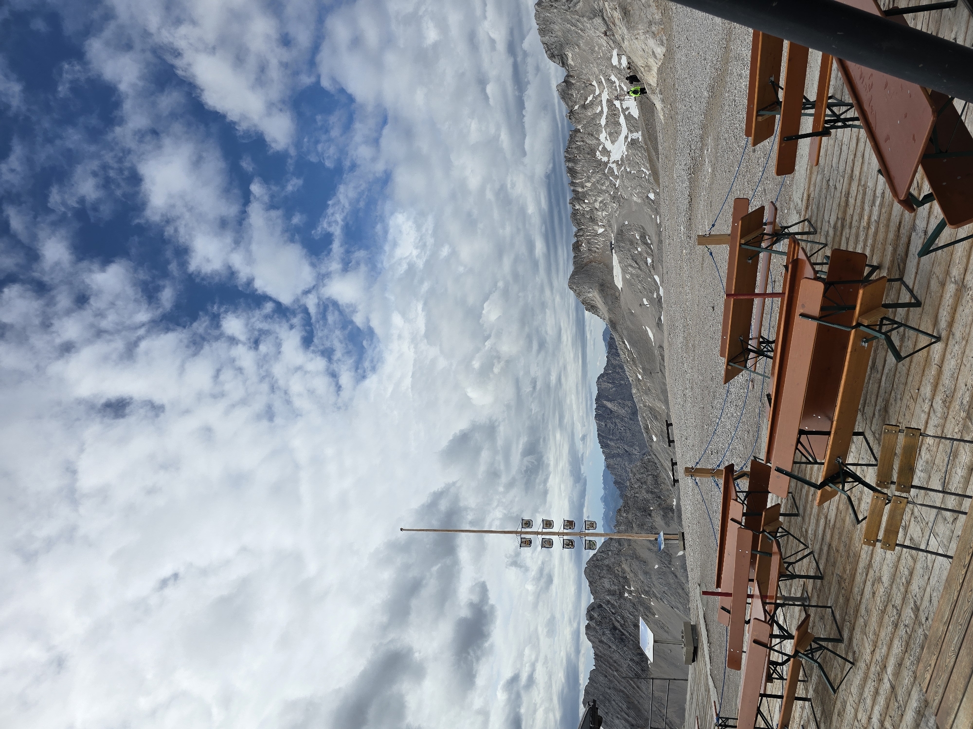 The image depicts an outdoor seating area with wooden tables and benches on a wooden platform. The platform is set against a backdrop of a rocky, mountainous landscape with patches of snow. The sky above is partly cloudy, with dramatic formations of white and gray clouds against a blue sky. In the mid-ground, a tall wooden pole stands with multiple small flags or banners attached to it. In the far background, more mountain ranges are visible, with some buildings or structures on a distant peak. Two individuals, dressed in outdoor clothing, are visible on the rocky terrain in the mid-ground, appearing to be walking. The scene appears to be a scenic viewpoint or an outdoor cafe at a high-altitude location. This location is likely the Zugspitze, Germany's highest peak, which is accessible from Garmisch-Partenkirchen.