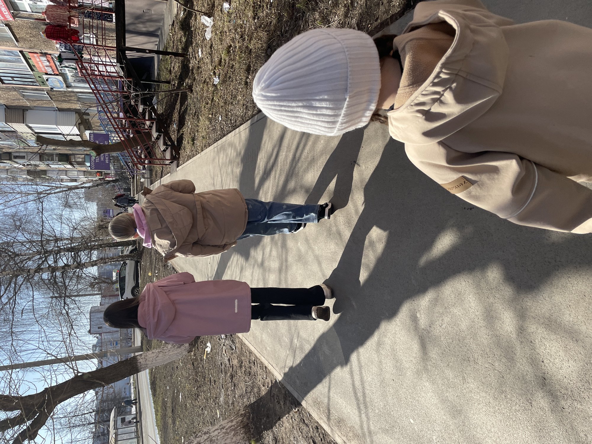 The image shows three children walking on a sidewalk in what appears to be an urban area. The first child, closest to the viewer, is wearing a beige jacket with a white knit hat. The second child is wearing a brown jacket and blue jeans. The third child is wearing a pink jacket and black pants. They are walking away from the camera. The sidewalk is concrete, and there is dry foliage on the side of the sidewalk. In the background, there are buildings, trees, and a metal staircase on the side of one of the buildings. Based on the context, the location is Izhevsk, Russia.