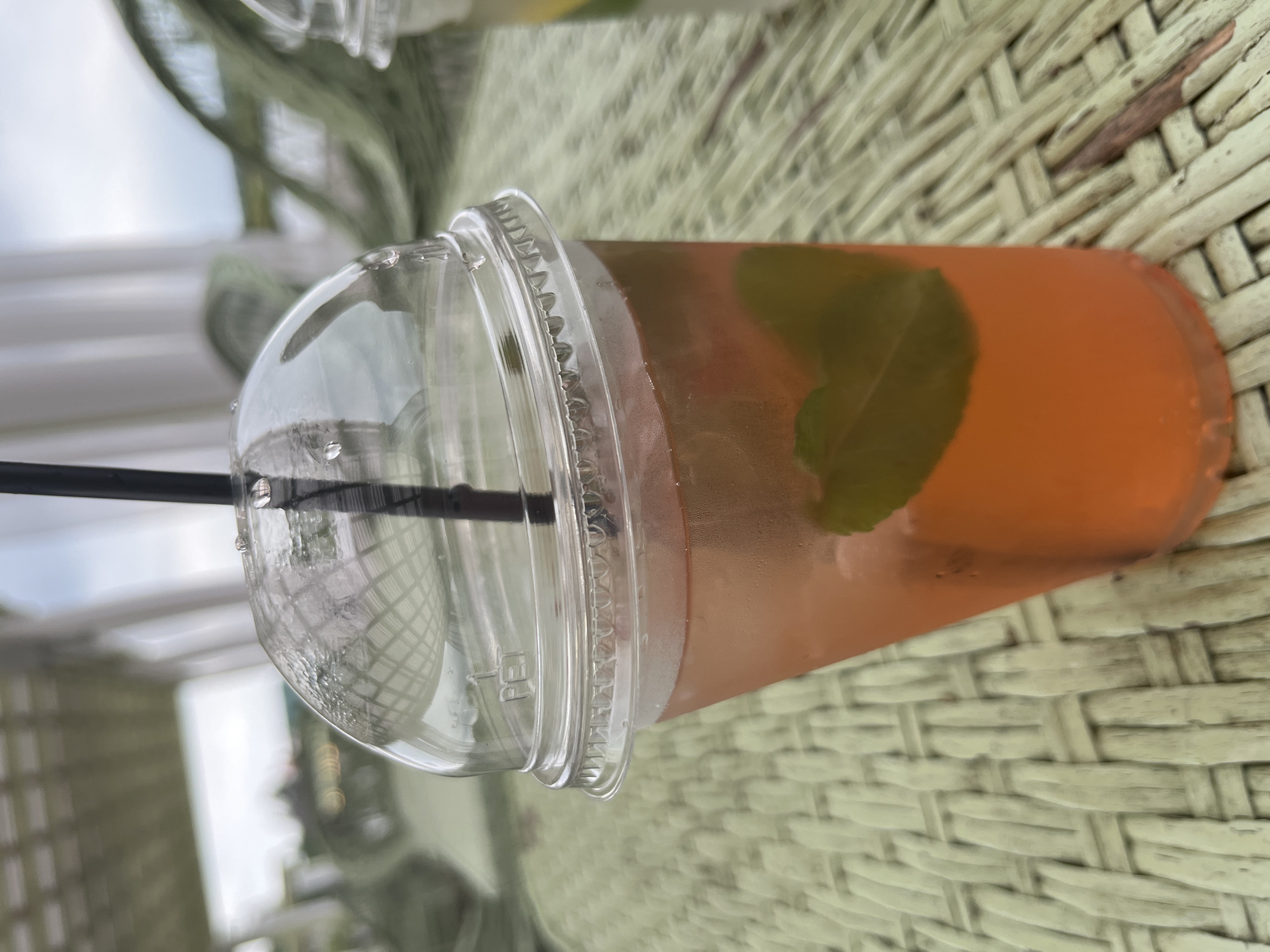 An outdoor scene captured in Severynivka, Ukraine, features a close-up of a clear plastic cup filled with a pinkish-orange beverage. A black straw extends from the top of the cup, which is covered by a domed plastic lid. Inside the drink, a fresh green leaf, likely mint, is visible, floating towards the bottom of the cup. The cup is placed on a textured, woven surface, possibly a table or placemat, with a pale green hue. In the background, out of focus, are hints of a covered outdoor seating area with white fabric drapes and a glimpse of a cloudy sky. Another partially visible plastic cup is present to the right of the main subject. The lighting suggests an overcast day.