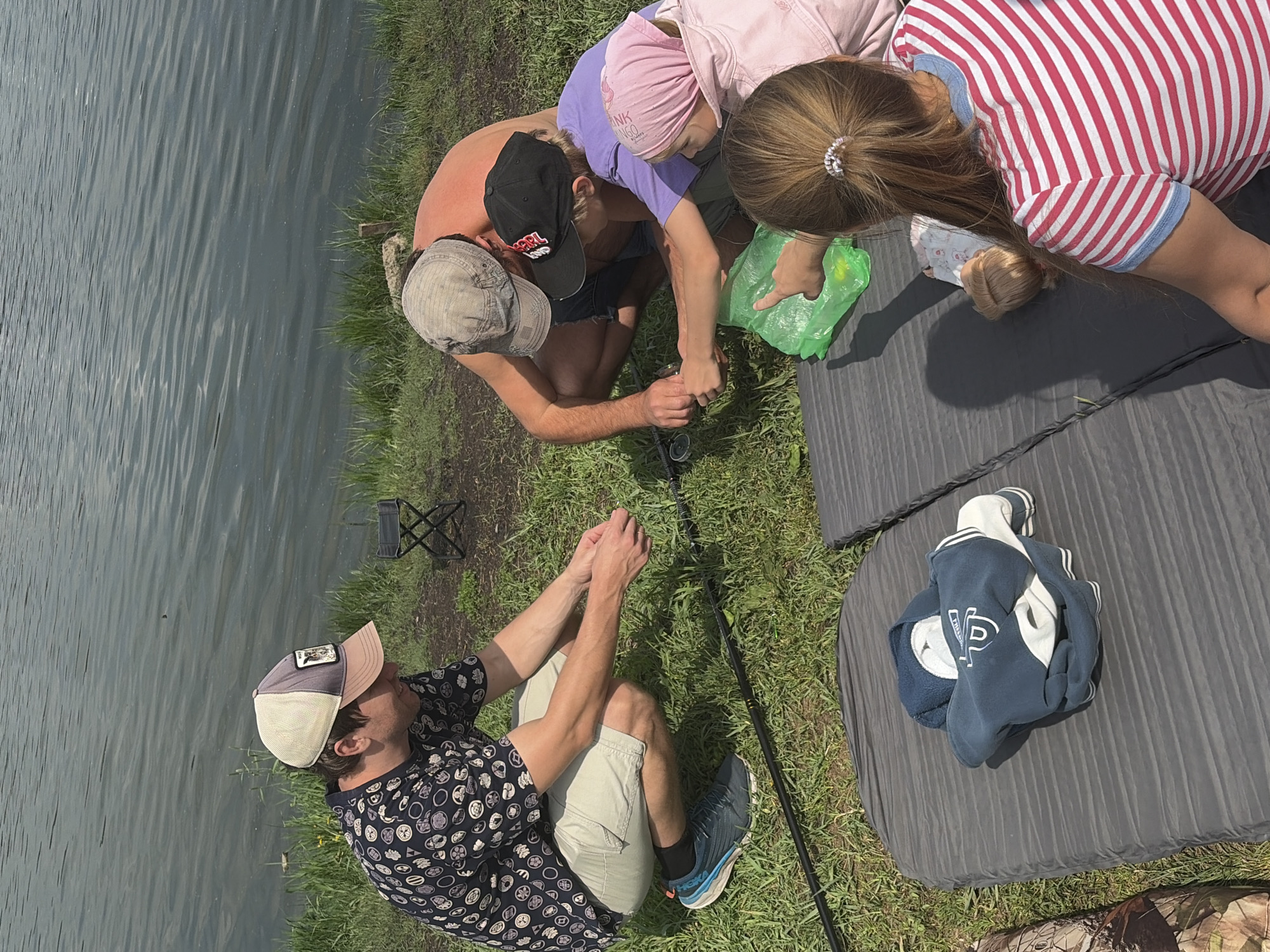 In the city of Minino, Russia, on a grassy bank beside a body of water, four individuals are engaged in an outdoor activity. A man wearing a patterned shirt and a baseball cap is crouching, facing away from the viewer, and appears to be handling fishing line. Beside him, another shirtless man in a baseball cap is also crouching. In front of them, two children are gathered. One child, wearing a purple shirt and a black baseball cap, is reaching towards a green plastic bag. A second child, wearing a pink headscarf and a pink jacket, is partially visible. In the foreground, two grey mats are laid out on the grass. On one mat, a blue and white hooded sweatshirt is folded. A person with long brown hair, wearing a pink and white striped shirt, is seated on the other mat, facing away from the camera and towards a doll. The water in the background appears calm, reflecting the overcast sky.