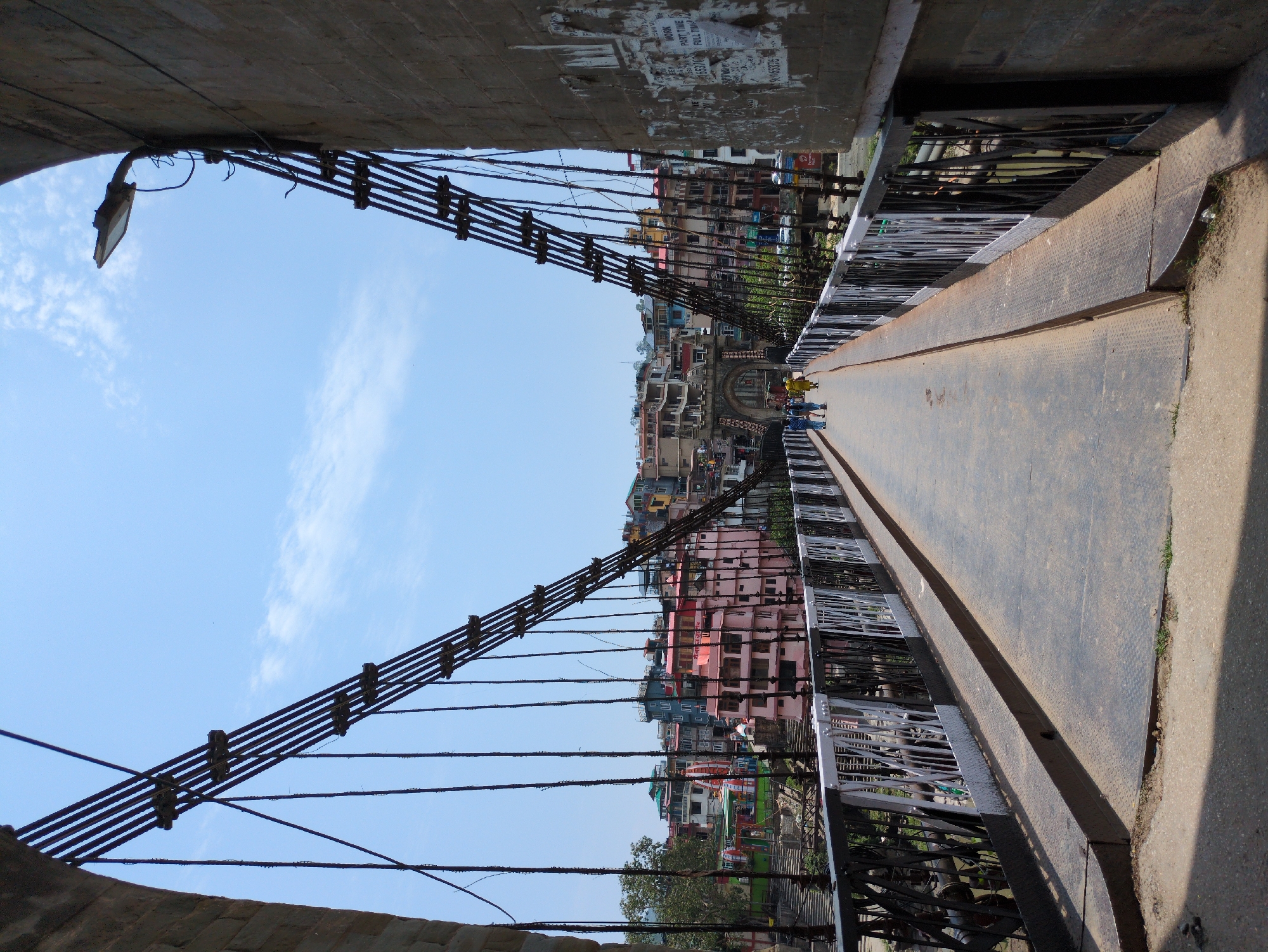 The image captures a view from a suspension bridge, likely in Mandi, India, looking towards a town built on a hillside. The bridge has a pedestrian walkway with a metal railing on either side. The railing is a combination of black and white metal bars, with a more intricate pattern of black metalwork visible beneath the white bars. Several people, appearing to be tourists, are walking across the bridge in the distance, with one person in a yellow dress being particularly prominent. The bridge's cables, made of thick metal wires, stretch upwards and outwards, anchored to stone structures on either side of the frame.
Beyond the bridge, the town is characterized by densely packed buildings in various colors, including pink, blue, and white, with multi-story structures reaching up the steep incline. Some buildings display architectural details like balconies and arched windows. In the foreground of the town, there is a visible temple with a white facade and colorful flags. The sky is a clear blue with a few wispy clouds, suggesting a sunny day. The overall scene depicts a functional suspension bridge serving as a pathway over a river or gorge, with a vibrant town nestled on the slopes beyond. The presence of advertising posters on the stone pillar to the right indicates a public space.