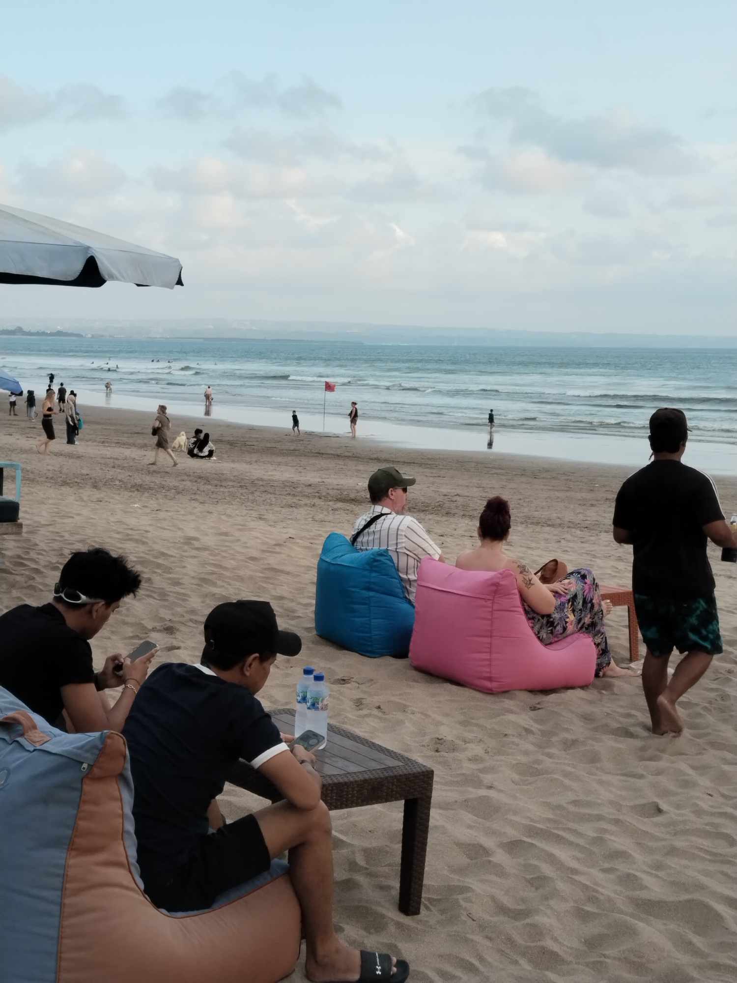 In Kuta, Indonesia, several people are relaxing on a sandy beach. In the foreground, two men are seated on beanbag chairs, looking at their phones. One man, wearing a black t-shirt and sunglasses, holds his phone. Next to him, a man in a dark blue t-shirt and a black baseball cap also uses his phone. A small table with two bottles of water sits between them. Further back, a group of people are seated on beanbag chairs facing the ocean. A woman in a colorful floral skirt and a man in a striped shirt are on blue and pink beanbag chairs. To their right, a man in a black t-shirt and patterned shorts stands, holding a bottle. In the background, the ocean stretches to the horizon under a cloudy sky, with a few figures visible near the water's edge. An umbrella provides shade on the left side of the image. The overall atmosphere is relaxed, with people enjoying the beach setting.