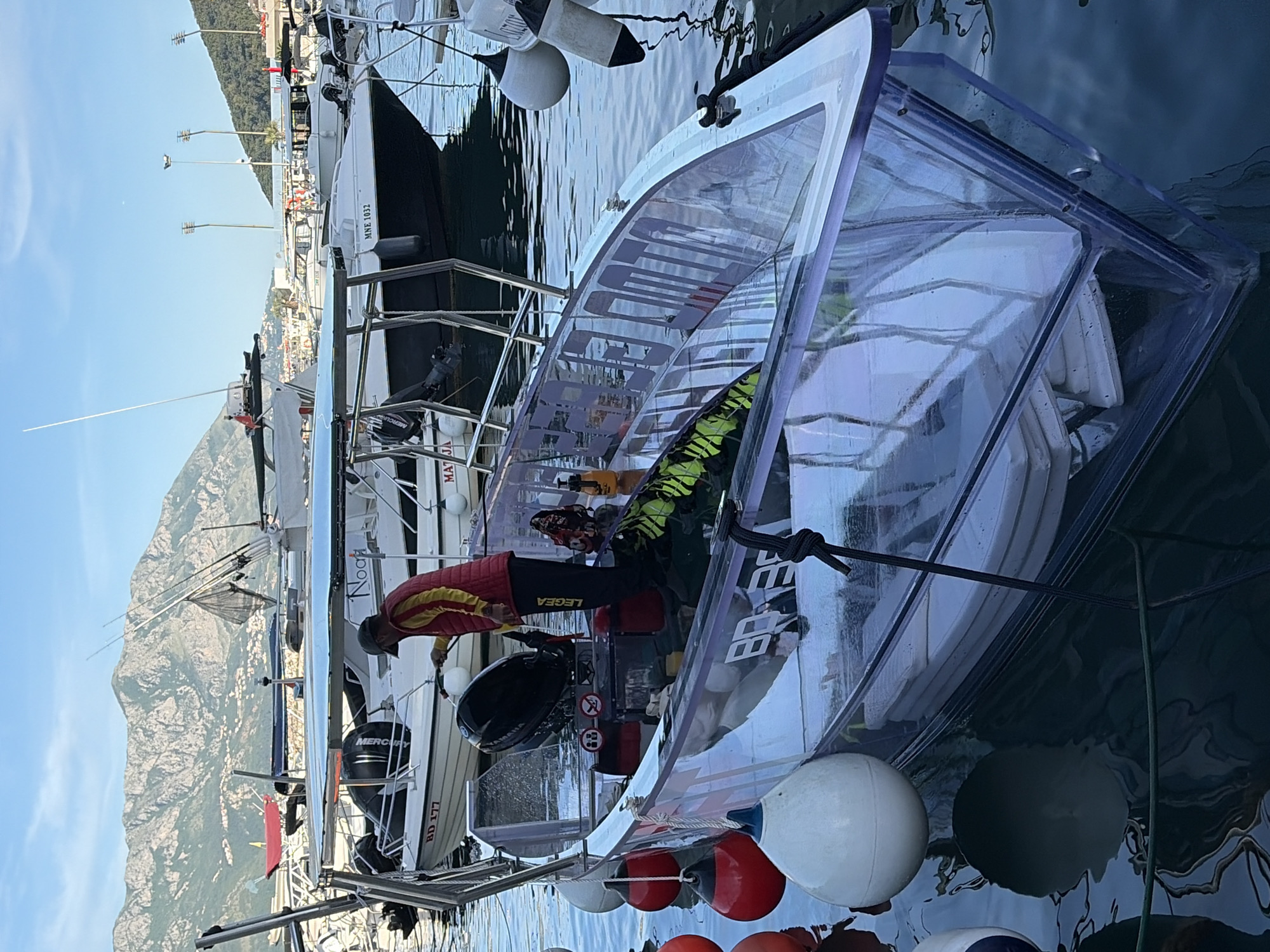 The image shows a marina in Budva, Montenegro, with a view of mountains in the background under a clear sky. Multiple boats are docked in the marina. The primary subject is a small boat featuring a transparent acrylic or similar clear plastic enclosure. A person wearing a red and yellow jacket, black leggings with the word "Legea" visible, and a dark hat is positioned next to the boat, appearing to be working on its motor. The motor is black and has the word "Yamaha" visible. A Mercury engine is attached to another boat behind the clear plastic vessel. A sign BD 177 is visible on another boat nearby. The dock has multiple white and red spherical buoys. The boat's hull has some letters on it. The water reflects the sky and surrounding objects. There are several streetlights on the waterfront in the distance.