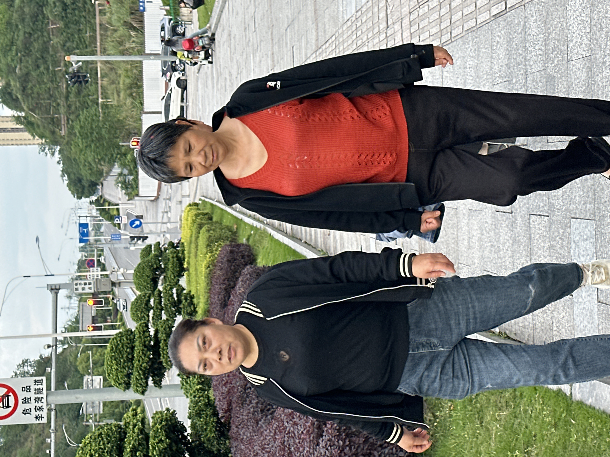 This image, rotated 90 degrees clockwise, depicts two women standing on a paved sidewalk next to a road in Jiangjin, China, during the daytime, likely under overcast conditions.
In the foreground, a woman wears a black tracksuit jacket with white stripes along the sleeves and body, blue jeans, and light-colored sneakers. She has short, dark hair and a neutral expression, looking towards the camera.
Behind her, a second woman wears a red patterned long-sleeved sweater under an open black jacket, black pants, and dark shoes. She also has short, dark hair and a gentle smile, looking slightly downwards.
The setting is an urban or suburban environment with a wide sidewalk made of square grey tiles. Adjacent to the sidewalk is a landscaped area featuring dense green and purplish-red bushes, along with trees. Beyond this greenery is a multi-lane road with traffic lights and streetlights. Cars and motorcycles are visible on the road and parked in the background. Hills covered in lush vegetation rise in the distance, with some buildings peeking through.
A notable detail is a red and white sign on the left side of the image (when correctly oriented) with a crossed-out vehicle symbol and Chinese characters that read "危险品 李家湾隧道" (Dangerous Goods Lijiawan Tunnel). Other blue and white road signs are also present. The overall atmosphere appears tranquil and mundane.