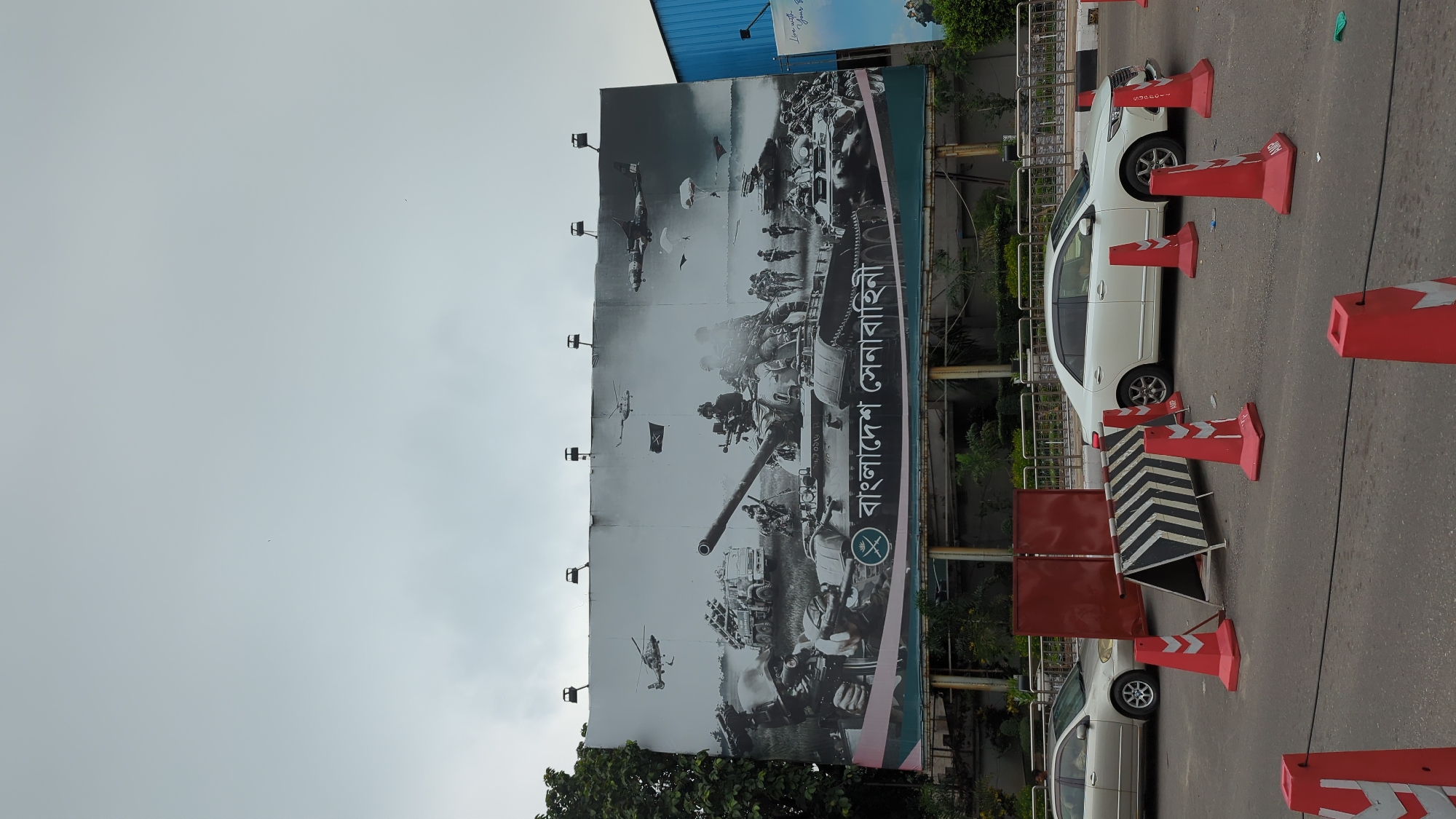 The image shows a street scene in Dhaka, Bangladesh. A large billboard dominates the view, displaying a black and white graphic of Bangladeshi military personnel and equipment, including tanks and helicopters. Text in Bengali, "বাংলাদেশ সেনাবাহিনী" (Bangladesh Sena Bahini), is prominently featured, indicating it's an advertisement or tribute to the Bangladesh Army.
Traffic cones and a black and white striped barrier partially obscure a white sedan parked along the roadside. The road is paved, and other vehicles are visible in the background. The sky is overcast. Trees are visible near the base of the billboard. The overall impression is that the photograph was taken on a busy urban street.
