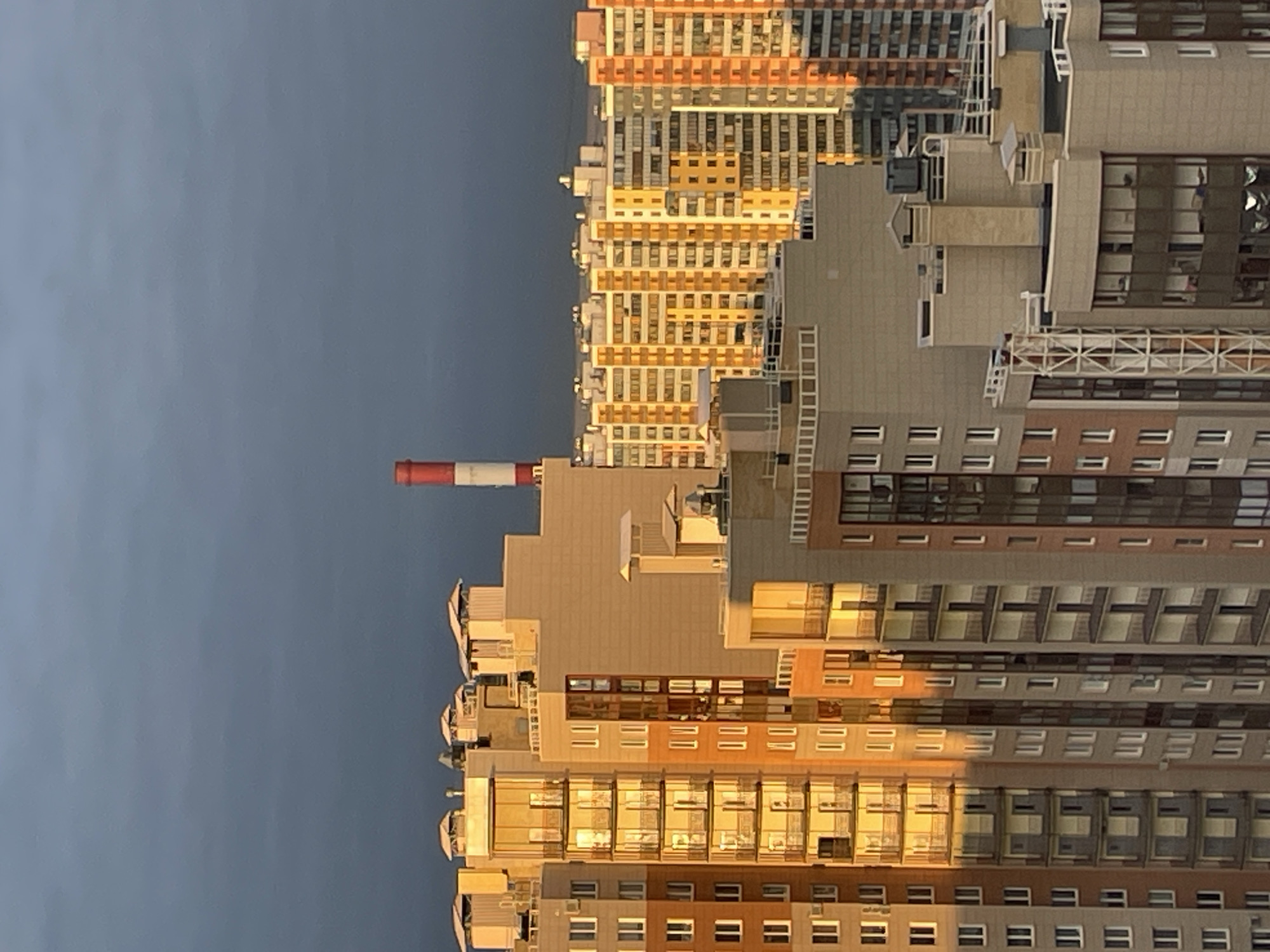 The image captures an urban landscape in Saint Petersburg, Russia, primarily featuring a dense cluster of modern high-rise buildings under a clear, pale grey-blue sky. The photograph appears to be taken during either sunrise or sunset, indicated by the warm, golden light illuminating the facades of the buildings from the left, casting long shadows and highlighting architectural details.
Numerous multi-story buildings dominate the lower and right portions of the frame. They exhibit varied architectural styles and colors, including shades of beige, grey, brown, and light orange/yellow. Many windows are visible, some reflecting the golden light, while others appear as dark openings. Balconies, ledges, and some exterior ladders or scaffolding structures are also discernible on the building surfaces. The buildings demonstrate a mix of textures and materials, creating a complex and detailed cityscape.
Standing tall against the sky, to the left of the main cluster of buildings, is a distinct industrial chimney or smokestack. It features alternating red and white horizontal stripes. No people or specific activities are visible within the frame, presenting a static view of the urban environment. The overall atmosphere is calm, with the soft, directional sunlight adding a dramatic quality to the otherwise structured urban scene. There is no visible text in the image.