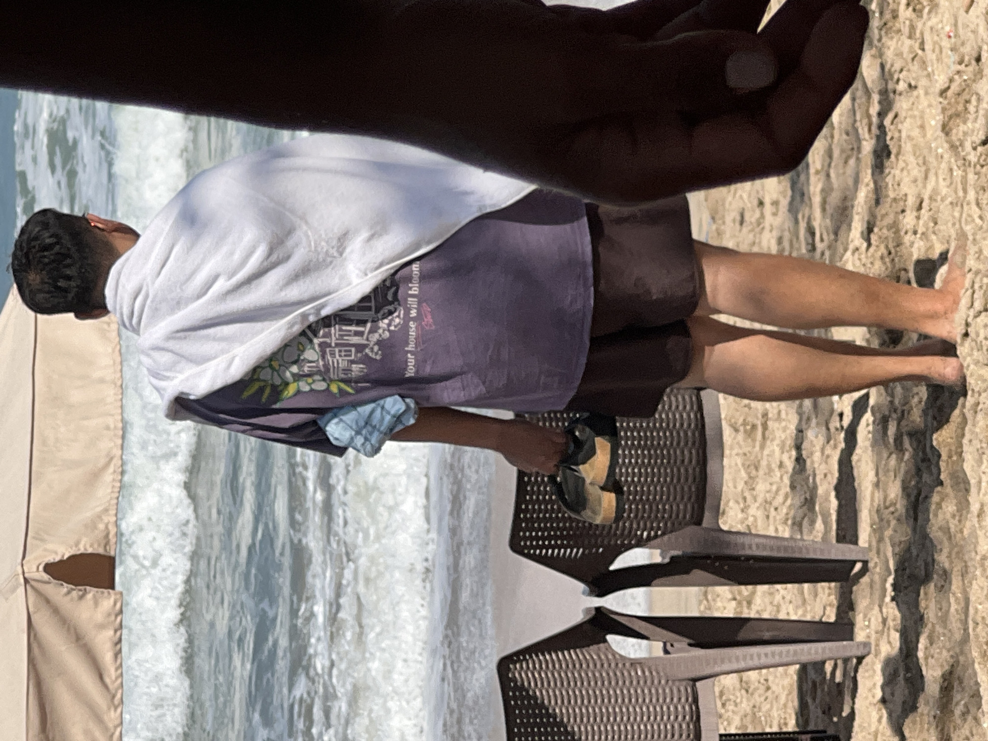 A man stands on a sandy beach in Kuta, Indonesia, holding a pair of sandals in his right hand. He is wearing a purple t-shirt, brown shorts, and a white towel draped over his shoulders. The ocean with waves is visible in the background, along with a light brown beach umbrella and two empty chairs. The man is facing away from the camera, looking towards the sea. Another person's hand is in the foreground on the right side of the image, partially obscuring the man's arm.