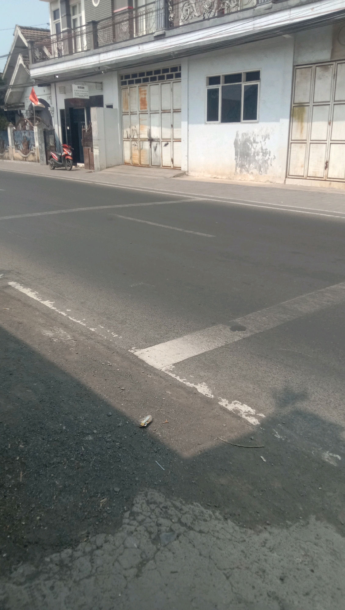 A street in Karang Anyar, Indonesia, features a building with white walls and large, segmented metal doors. A red motorcycle is parked in front of the building. The street is paved and has white traffic markings. The scene appears to be captured during the daytime under sunlight, casting shadows on the road.