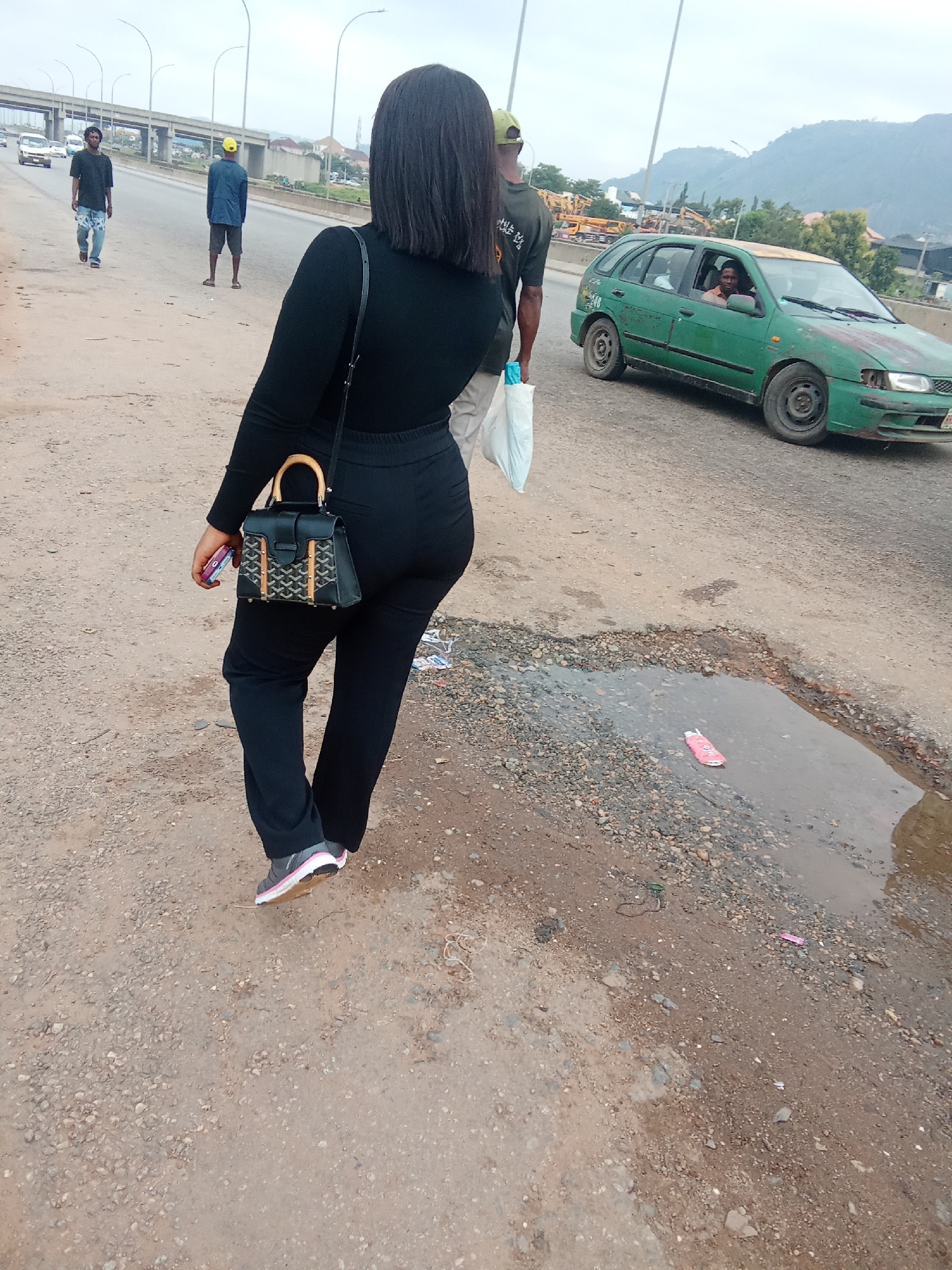 This media file depicts a street scene in Kagini, Nigeria, on an overcast day. The main subject is a woman walking away from the camera, wearing a black long-sleeved shirt, black pants, and athletic shoes with pink accents. She is carrying a small, black handbag with a bamboo handle and a decorative pattern on the front, and also holding a pink phone in her left hand.
In the background, two men are walking on the side of the road. One man wears a dark t-shirt and patterned shorts, while the other wears a blue shirt and dark shorts, with a yellow cap. Further in the background, a green hatchback car, possibly a taxi, is driving by. A man is visible in the driver's seat. The road surface is rough and unpaved, with several potholes and a large puddle of murky water, containing a pink object, likely litter. The scene is set against a backdrop of a highway overpass and mountainous terrain, suggesting an urban or semi-urban environment. The overall atmosphere is ordinary and appears to be a typical day with no specific event taking place, other than people going about their daily activities. No discernible text is visible in the image.