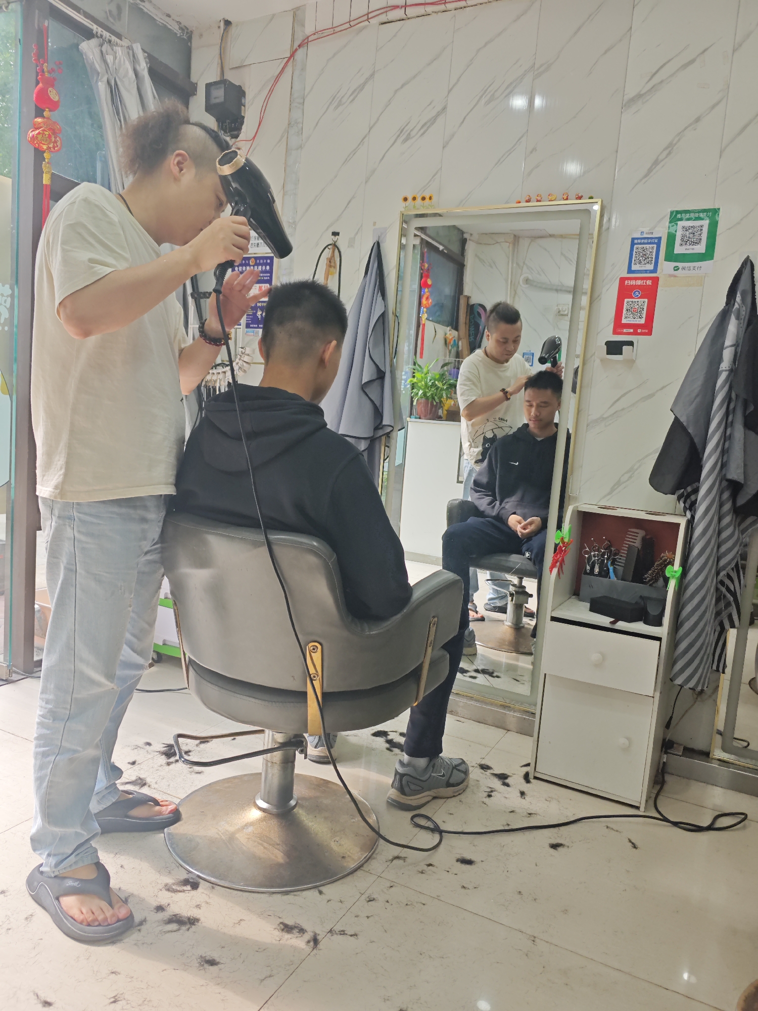 In a barbershop located in the Chongqing High-tech Zone, China, a hairdresser is blow-drying the hair of a seated customer. The customer, wearing a black hooded sweatshirt and dark pants, is seated in a grey barber chair, facing away from the camera. The hairdresser, in a white t-shirt and light blue jeans, stands behind the customer, holding a black hairdryer and aiming it at the customer's head. The floor around the chair is scattered with cut hair. A large mirror reflects the scene, showing another man in the background also blow-drying someone's hair. The walls of the barbershop are tiled in a white marble pattern. Various items are visible, including a white cabinet with drawers and shelves containing hairdressing tools, as well as QR codes for payment methods affixed to the wall. A black power cord snakes across the floor.