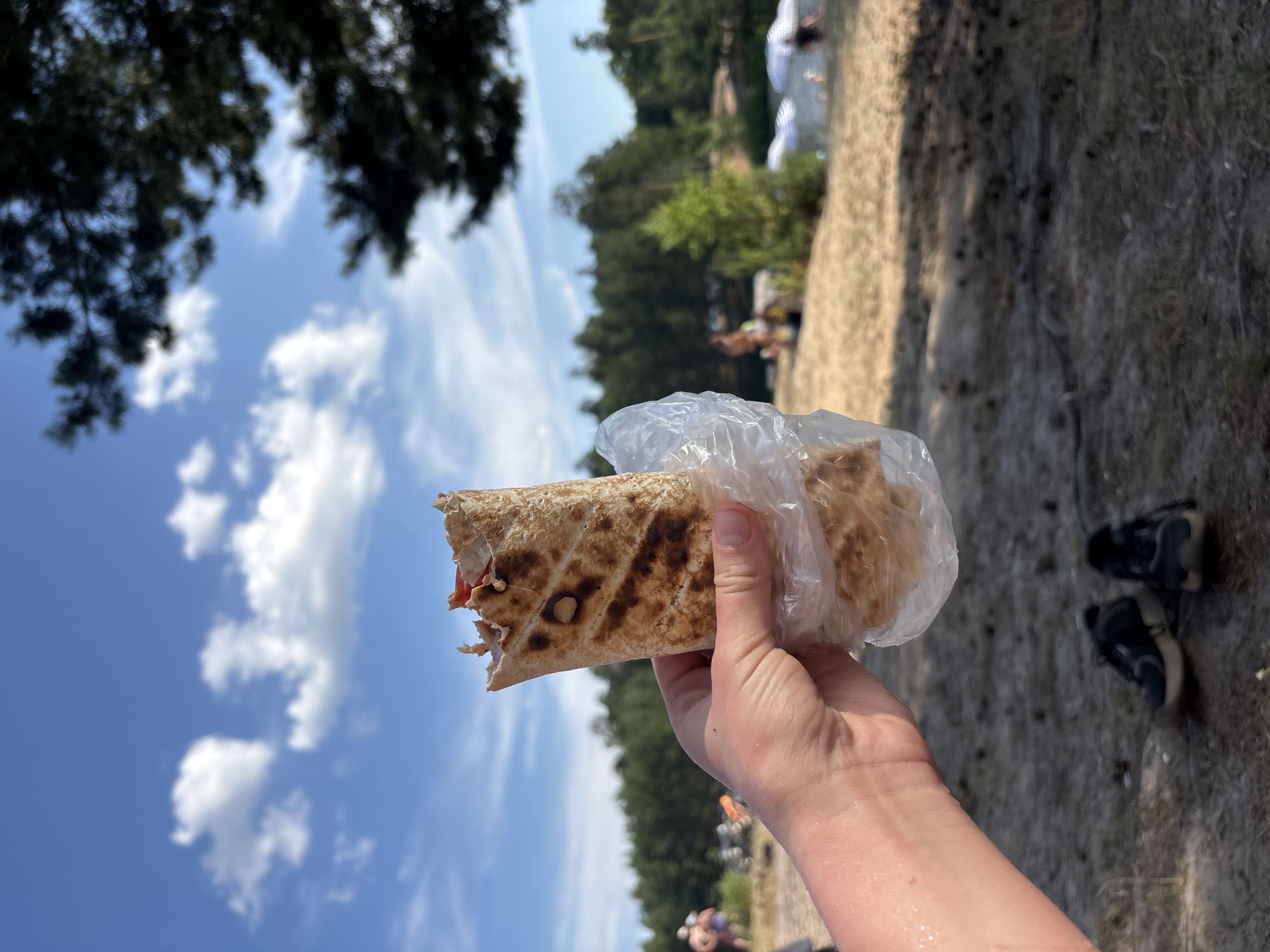 A person holds a partially eaten shawarma wrapped in plastic in their left hand. The shawarma has grill marks and visible filling. The background is a sandy area with sparse vegetation, and a body of water with beach umbrellas and people in the distance. A line of trees forms the horizon. The sky is blue with scattered white clouds. A pair of dark athletic shoes are on the ground in the foreground. The image appears to be taken on a sunny day.