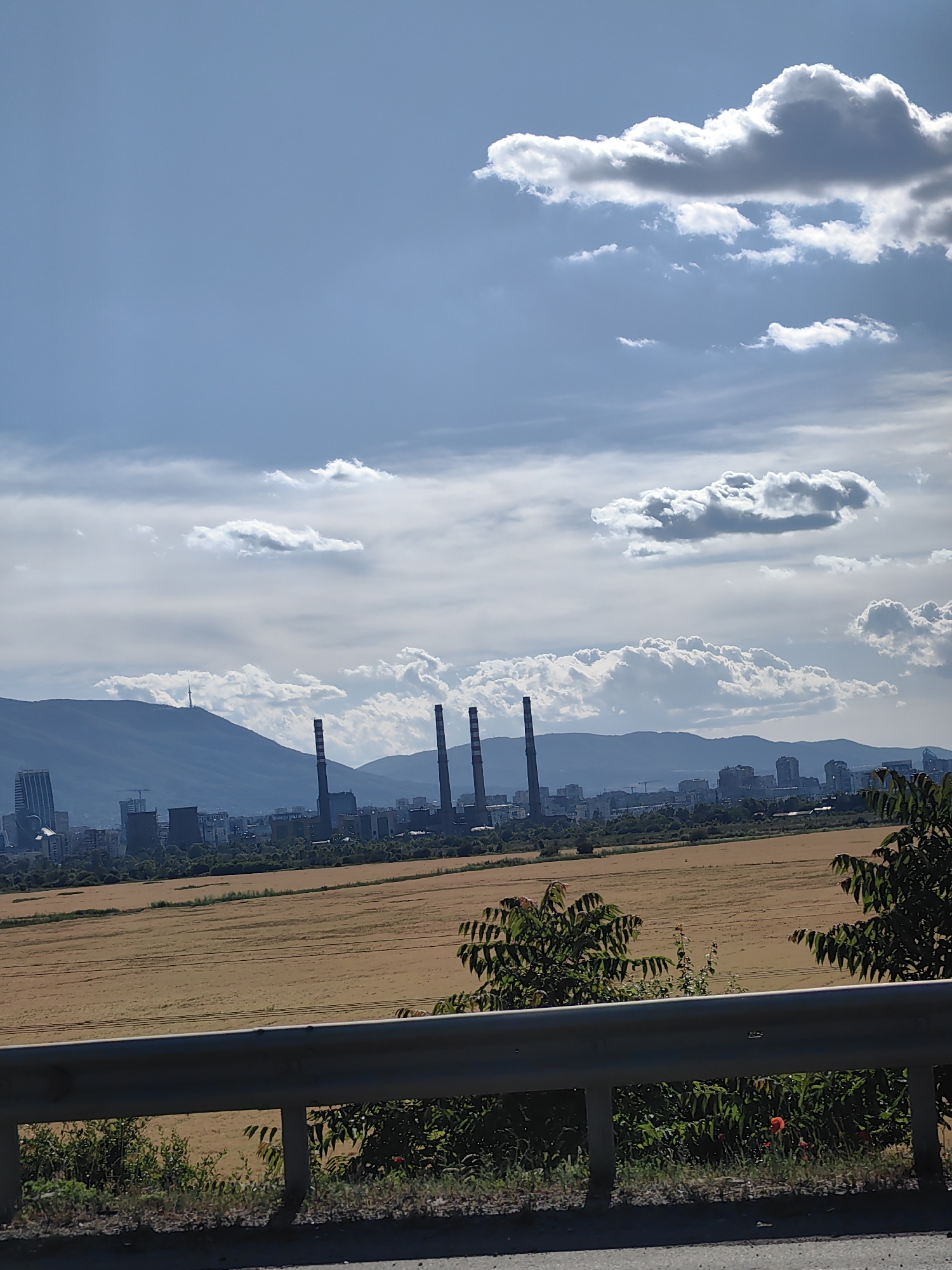 The image depicts a panoramic view of Kazichene, Bulgaria, under a partly cloudy sky. In the foreground, a metal guardrail separates the viewer from a field of dry, golden crops. Behind the field, a city skyline is visible, with numerous buildings and industrial structures, including several tall smokestacks from a power plant. In the distance, a mountain range forms the backdrop, with a prominent television tower on one of the peaks. The overall scene suggests an industrial or semi-urban area, with elements of nature present in the fields and mountains. There are no people or animals visible in the image.
