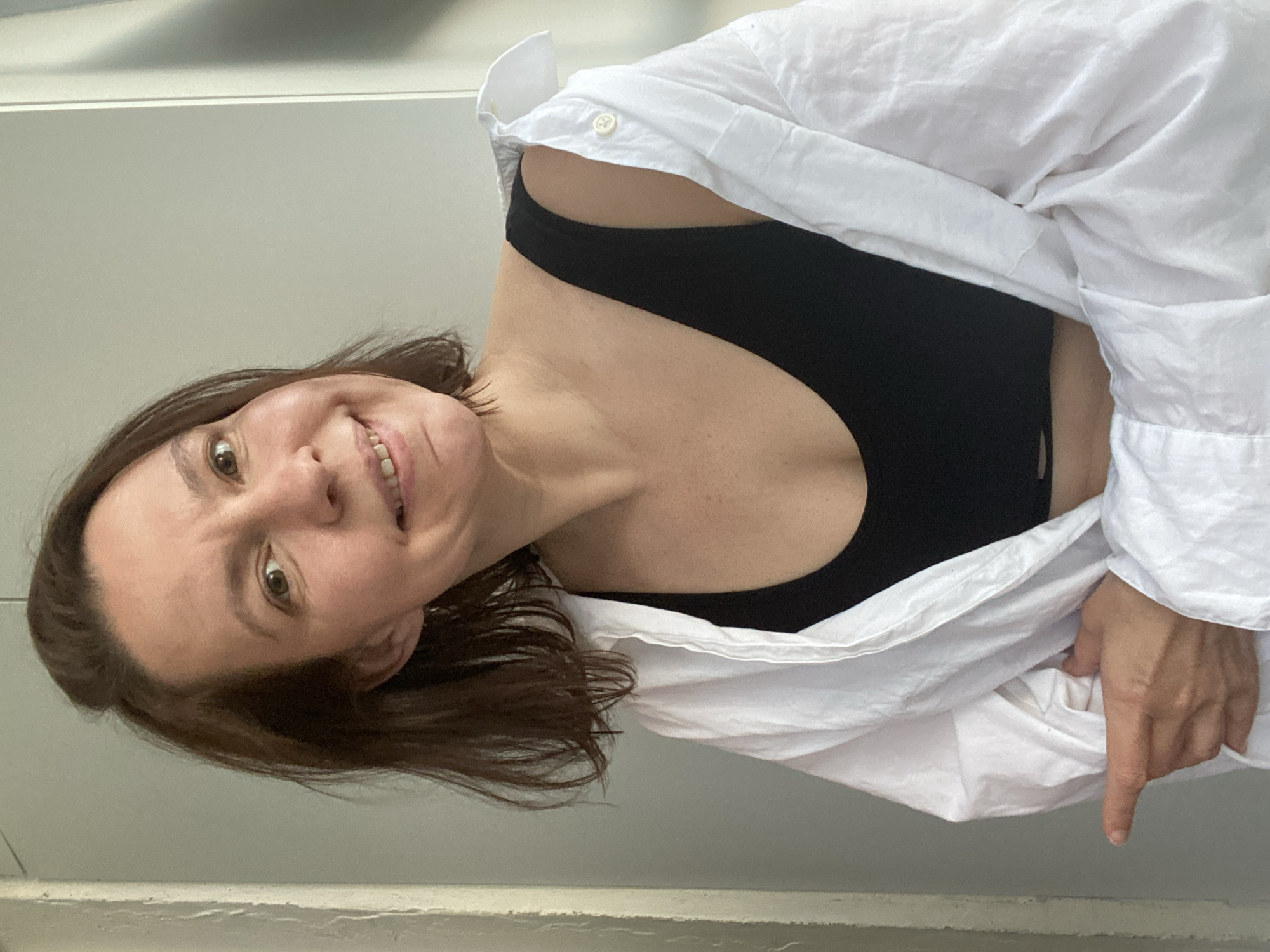 A woman is indoors, wearing a black tank top and a loosely draped white button-down shirt. She is smiling and looking towards the camera. Her hair is shoulder-length and brown. Her hand is visible, pointing downwards. The background is a light gray wall. The location information is unavailable.