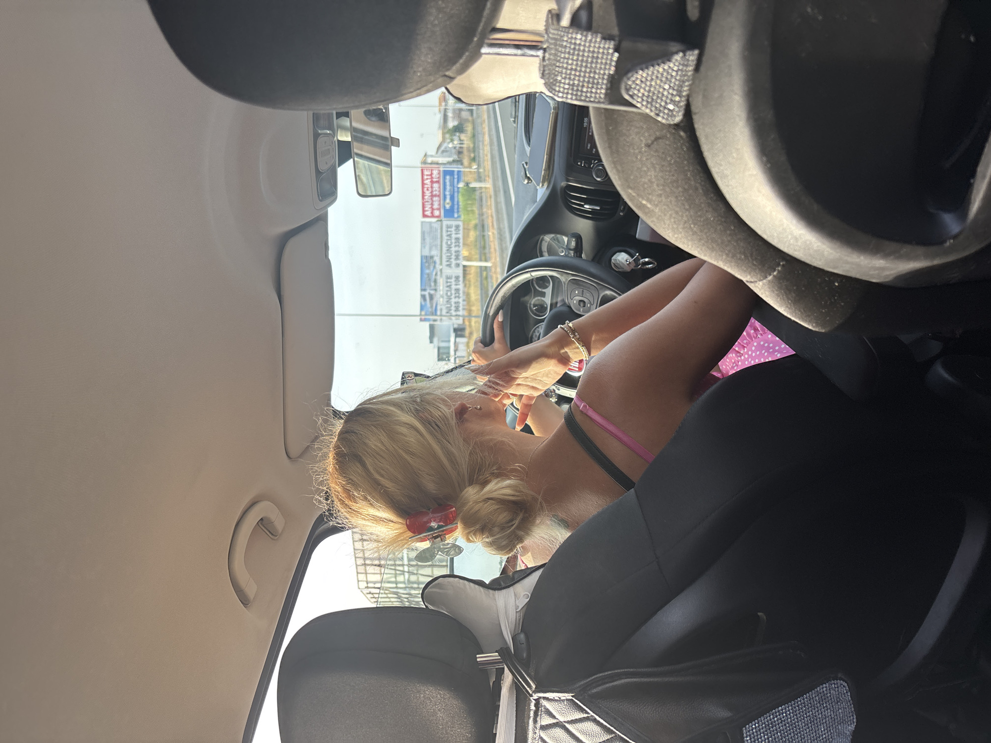 A woman with blonde hair pulled back in a bun, accessorized with a red and silver butterfly hair clip, is driving a car on a sunny day. She is wearing a pink top with thin straps and a pink dress with white polka dots. She has a bracelet on her left wrist and a ring on her right ring finger. The car's interior is visible, including the steering wheel, dashboard with a phone on it, and the headrests of the seats. Outside the car, billboards for "ANUNCIATE" with phone numbers are visible along the road, suggesting a commercial or urban setting. The scene appears to be a highway or main road in Urbanización Cabo Roig, Spain.