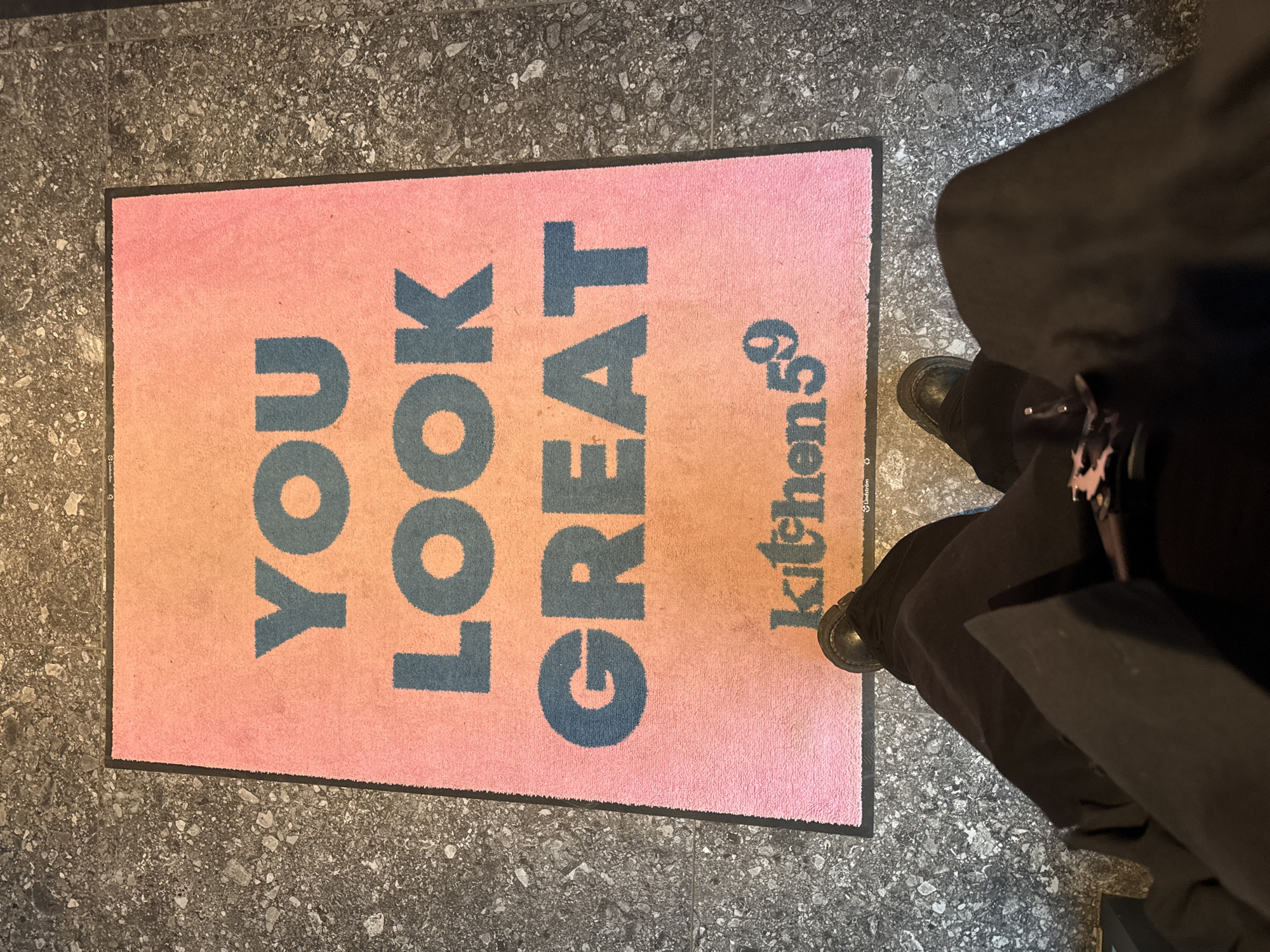 The image shows a doormat with the words "YOU LOOK GREAT kitchen59" in blue lettering against a pink background. The mat is rectangular and placed on a grey, speckled floor. A person is standing on the mat, wearing black boots and dark clothing. The location is not discernible from the image provided, and cannot be determined to be Sofia, Bulgaria.