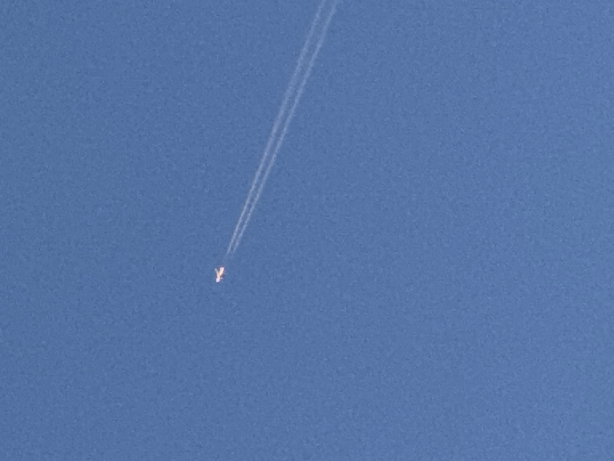 The image shows a clear blue sky with a single airplane flying at a high altitude. The airplane is leaving a prominent contrail behind it, which consists of two distinct white lines. The airplane itself appears as a small, bright object against the vast expanse of the sky. There are no visible buildings, landmarks, or any other contextual elements that would suggest a specific location within London, United Kingdom. The focus of the image is solely on the airplane and its contrail in an otherwise featureless sky.