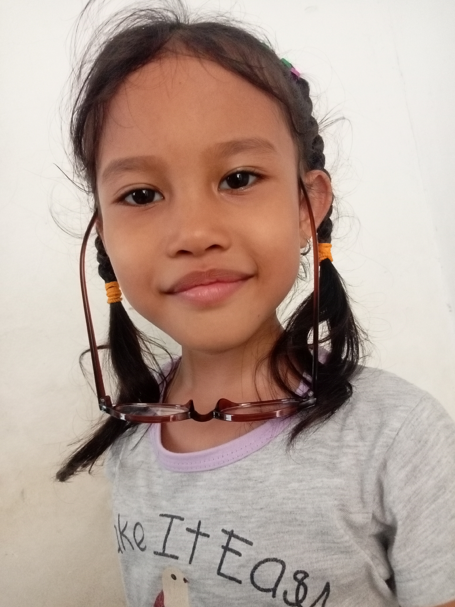 A young girl with dark hair in two braids, secured with orange bands, poses for a selfie. She is wearing a grey t-shirt with a light purple trim around the neckline. A pair of brown-rimmed glasses are resting on her braids, hanging over her ears. The background is a plain white wall. The image appears to be taken indoors, likely in a residential setting, possibly in Batam, Indonesia, given the context. The girl is looking directly at the camera with a slight smile. The lighting is natural and even, suggesting the photo was taken during the day. The overall tone of the image is casual and personal.