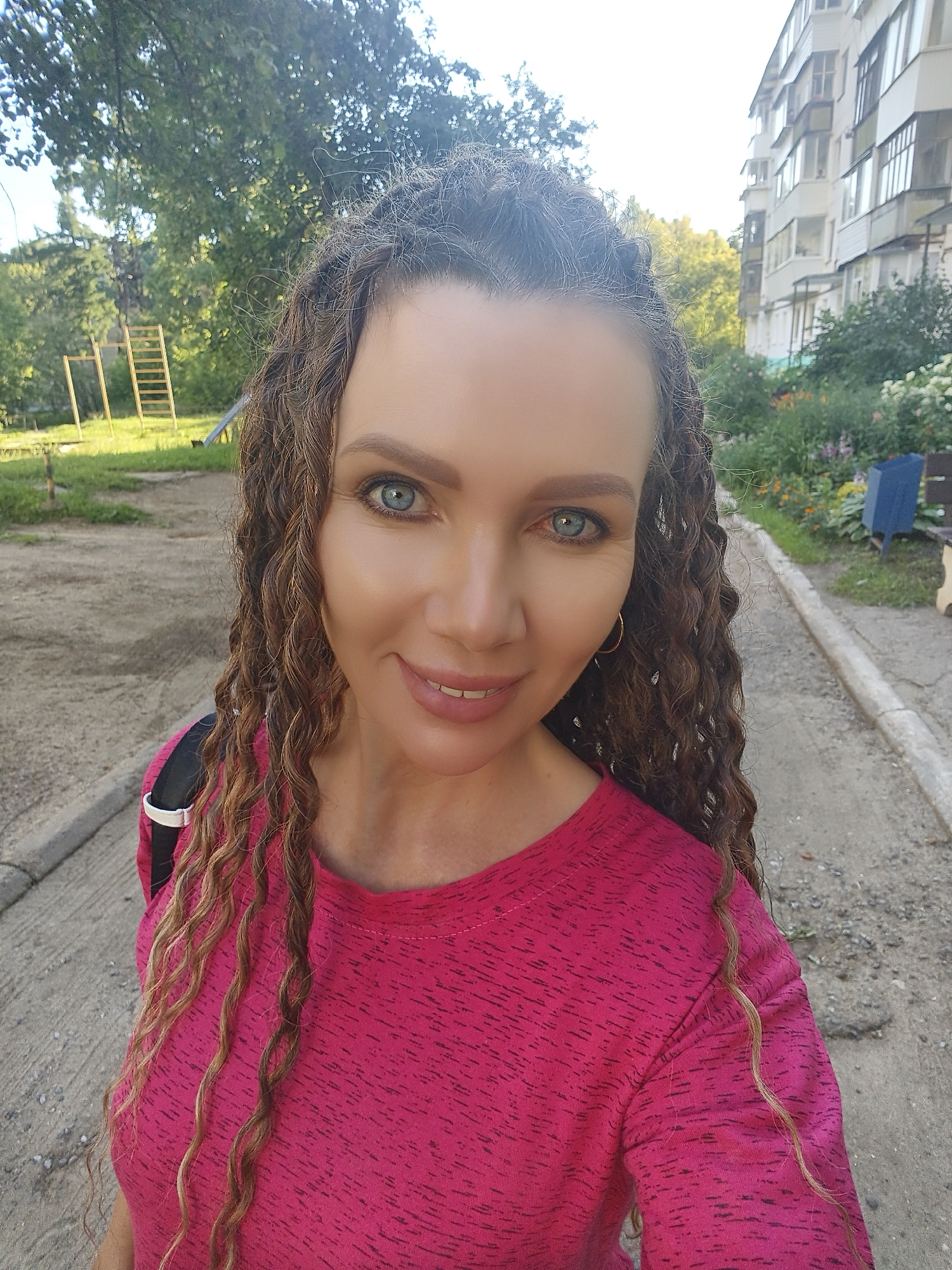 A woman with light skin, blue eyes, and long, curly brown hair is taking a selfie outdoors. She is wearing a bright pink, crew-neck t-shirt with a black backpack strap visible over her shoulder. The woman is smiling, showing her teeth. Her hair is styled in loose curls that fall down past her shoulders. She is standing on a paved surface, possibly a sidewalk, with a gravelly area to her left. In the background, there is a patch of grass with a children's playground structure, including a ladder. To the right of the woman, there is a building with multiple stories and balconies, and a flower bed with colorful blooms. The sky is visible in the upper portion of the image, appearing bright and possibly overcast or hazy.