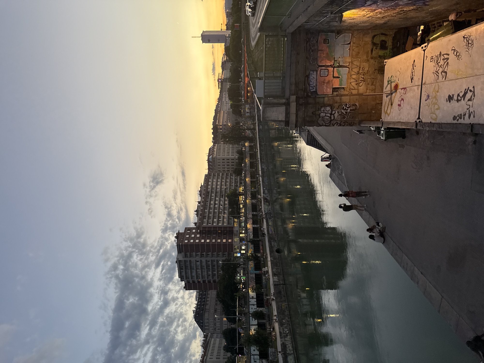 The image captures a waterfront scene in Vienna, Austria, during what appears to be sunset or dusk. The sky transitions from a pale blue to a warm yellow-orange hue, indicating the evening light. A canal or river runs alongside a paved walkway, reflecting the lights from the buildings and the sky.
On the left side of the image, a dense cityscape of apartment buildings rises, their windows illuminated, suggesting the time is evening. The architecture appears to be a mix of older and more modern styles. Trees line the walkway in front of the buildings, adding a touch of greenery.
The walkway itself is populated by several individuals. Some are standing, seemingly observing the surroundings or talking to each other. A few are seated, perhaps resting or enjoying the ambiance. The mood appears relaxed and peaceful.
On the right side of the canal, a concrete structure, possibly a bridge abutment or retaining wall, is heavily adorned with graffiti. The graffiti features various tags and artistic designs in multiple colors. A large, pale concrete block structure, also covered in graffiti, is visible.
The water is calm, with gentle reflections creating an impressionistic effect. The overall impression is one of an urban environment transitioning into evening, with a mix of residential areas, public spaces, and artistic expression. The time of day is evident from the lighting conditions, and the location is suggested by the urban architecture.