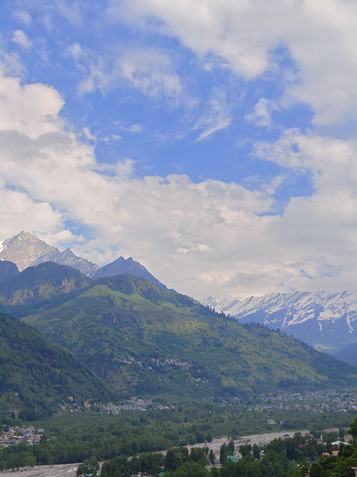 The image depicts a panoramic view of the Himalayan mountains, specifically the region around Vashisht, India. In the foreground, a lush green valley is dotted with a settlement of multi-story buildings, a stark contrast to the natural landscape. Dense green trees cover the lower slopes of the mountains, which rise dramatically in the midground. These midground mountains are covered in vibrant green vegetation, with some areas appearing as terraced fields or natural grasslands. Higher up, the mountain peaks are rugged and jagged, with patches of snow still visible on the highest elevations, indicating a mountainous, alpine environment. The sky above is a vibrant blue, adorned with a scattering of white, puffy clouds that create a dynamic and atmospheric backdrop. The overall scene is one of serene natural beauty, with human habitation nestled harmoniously within the grandeur of the Himalayas.
