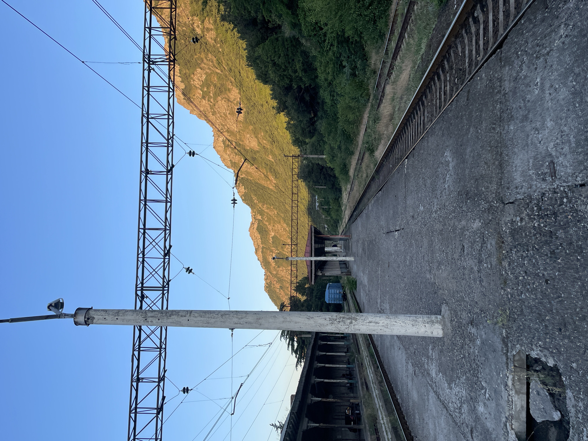 This is a vertical, slightly low-angle shot of a train platform and tracks next to a steep, forested mountain. The scene is set in Mtskheta, Georgia. The time of day appears to be late afternoon or early evening, as the sun casts a warm, golden light on the mountain face, illuminating the rugged terrain and the dense green foliage. The sky is a clear, bright blue, suggesting fair weather.
In the foreground, the concrete platform is worn and weathered, showing signs of age and damage, with a large hole revealing some debris underneath. A tall, white pole with a lamp fixture stands prominently on the platform, casting a shadow. To the left of the platform, a lattice-like metal structure supports overhead electrical wires, typical of a railway line. The tracks themselves are visible, running parallel to the platform and disappearing into the distance.
In the middle ground, there is a small, covered structure that resembles a bus stop or a small station building, with a red roof. A blue portable toilet is parked nearby. The mountain rises steeply behind the tracks and the building, its slopes covered in trees that transition from dark green to a sun-drenched golden hue on the upper parts.
The overall mood of the image is one of quiet desolation and natural beauty. The abandoned-looking platform and the vastness of the mountain suggest a sense of remoteness. There is no visible text in the image.