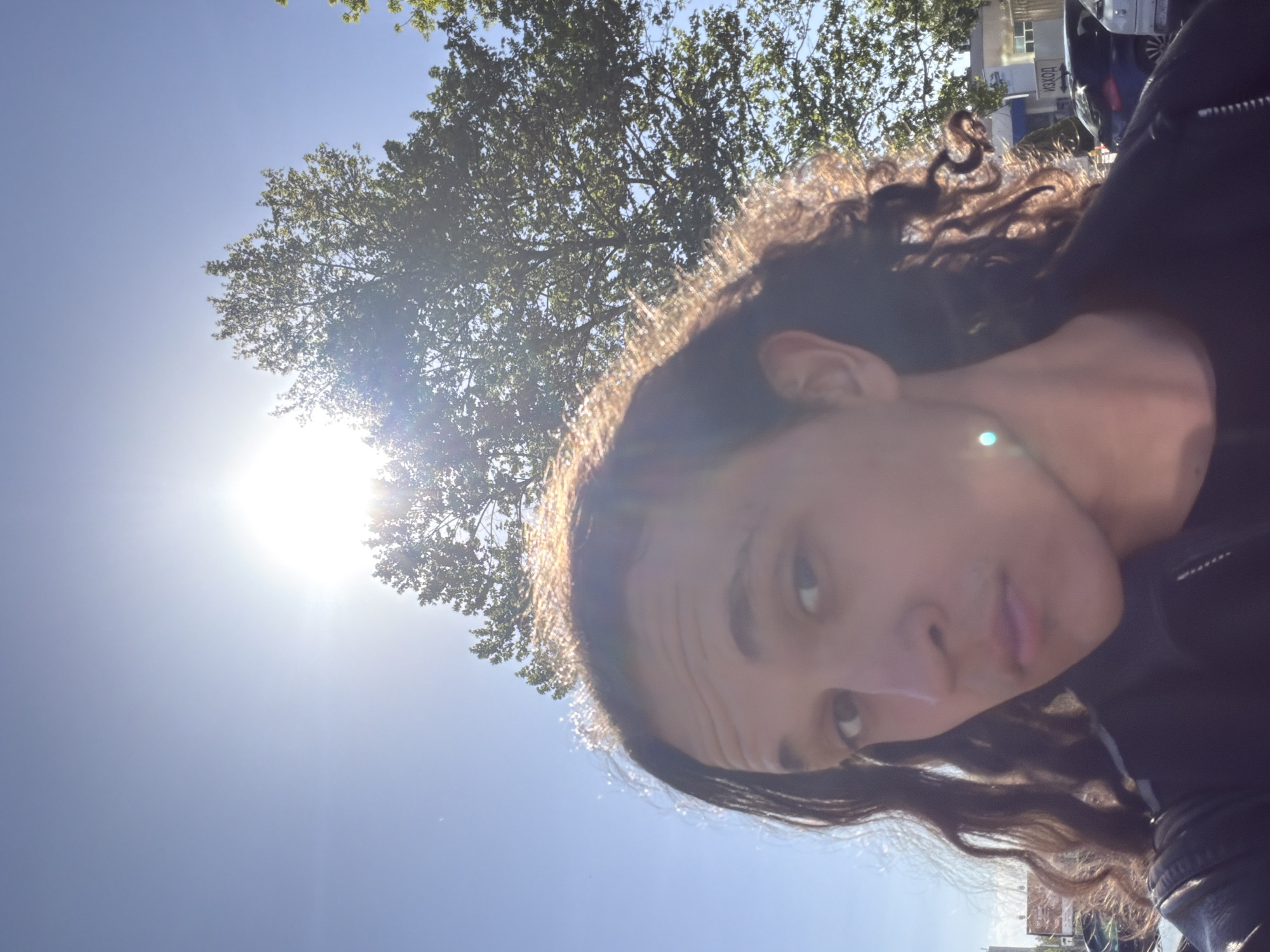 The image is a bright, sun-drenched outdoor selfie, rotated approximately 90 degrees clockwise. The primary subject is a young person, likely female, with dark, curly hair illuminated by strong backlight from the sun. They wear a dark jacket and look directly towards the camera with a neutral expression.
The background is dominated by a clear blue sky and abundant green foliage from large trees, suggesting a natural or park-like setting within an urban environment. The sun is a powerful light source, creating significant lens flare and strong highlights on the subject's hair.
In the upper right portion of the original image, behind the subject's head, urban elements are visible: a building with windows and the side of a vehicle, possibly a car or truck. Notably, Cyrillic text reading "ИЗХОД" (IZKHOD), meaning "EXIT" in Bulgarian, is visible on the building, consistent with the specified location of Plovdiv, Bulgaria.
The scene depicts a sunny daytime, likely midday or early afternoon given the intensity of the light. The weather is clear and bright. No specific activity is shown beyond the act of taking a photograph outdoors.