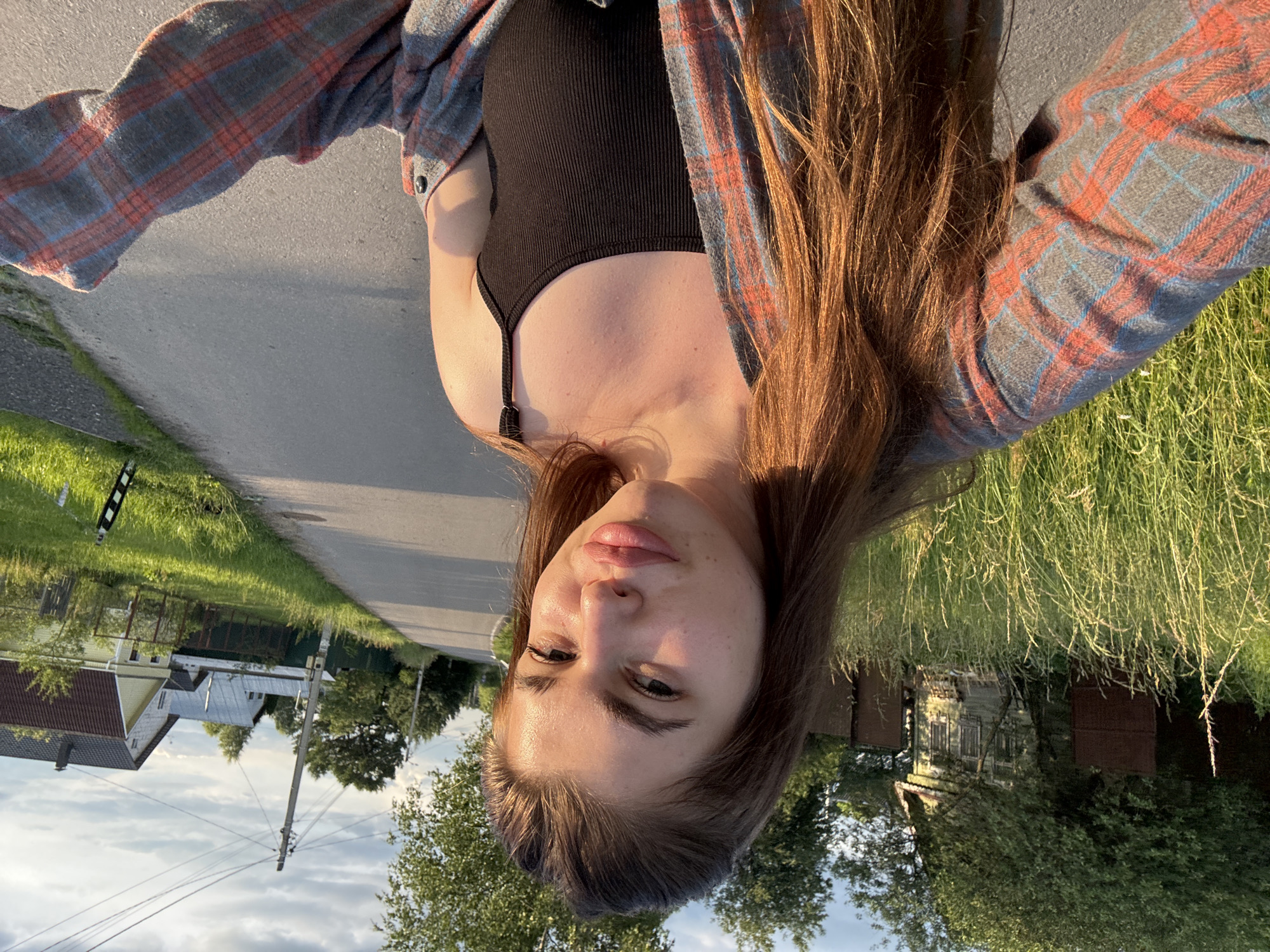 A young woman with long, brown hair is captured in a selfie, outdoors on a paved road. She is wearing a black tank top and an open plaid shirt in shades of grey, red, and blue. Her hair is styled naturally and falls around her shoulders. She is looking directly at the camera with a neutral expression, her lips slightly parted. The background suggests a rural setting with houses and trees visible on either side of the road. The sun is low in the sky, casting long shadows and illuminating the scene with warm, golden light, indicating it is likely late afternoon or early evening. The sky is partly cloudy. The image is oriented sideways.